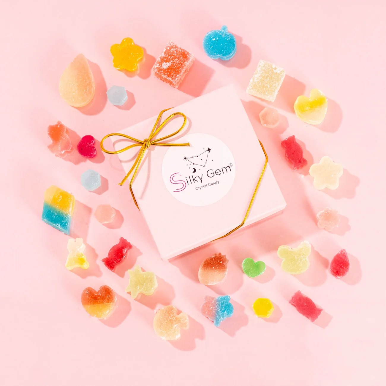 A pink box labeled 'Silky Gem Crystal Candy' is on a pink background, surrounded by colorful gummy candies in shapes like hearts, stars, flowers, and bears.
