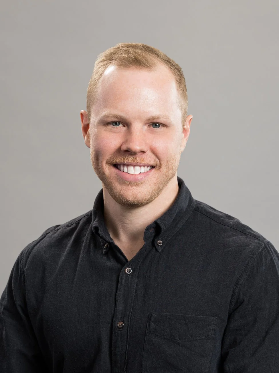 Headshot of a man with short, red hair and a beard, smiling, wearing a black button-up shirt against a gray background.