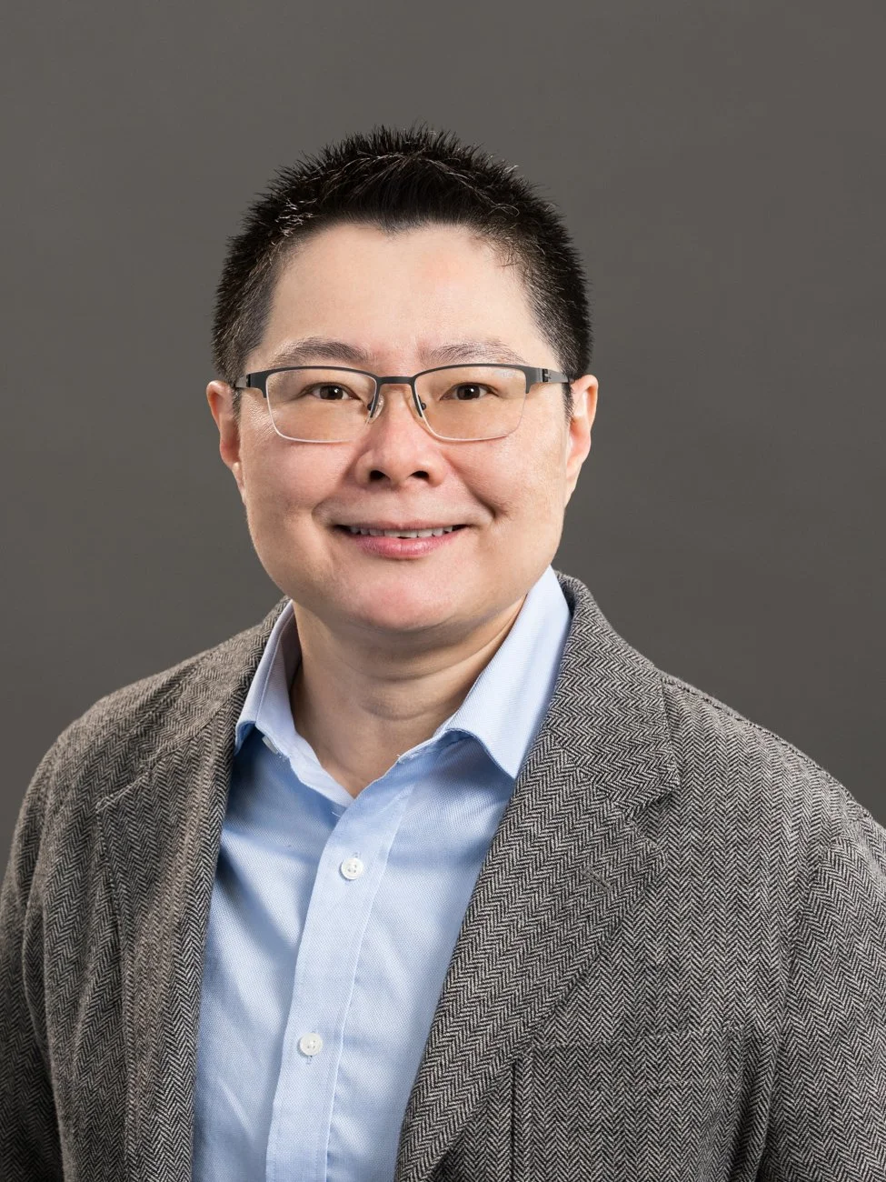 Headshot of an Asian man with black hair, glasses, smiling, wearing a blue shirt and gray blazer against a gray background.