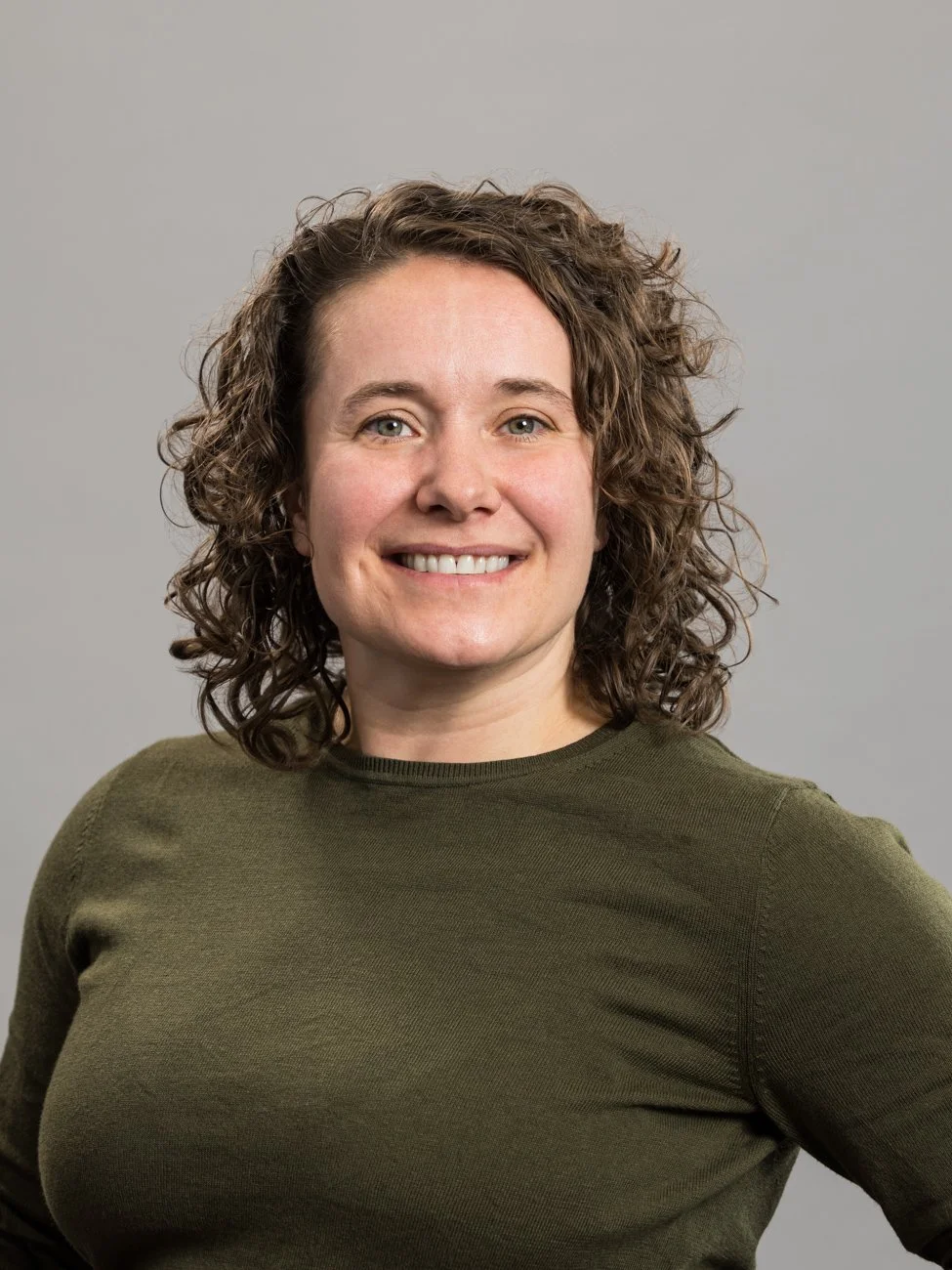 A woman with curly brown hair wearing a dark green shirt, smiling at the camera, against a gray background.