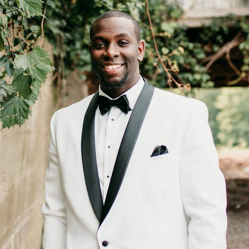A man smiling in a white tuxedo with black lapels and bow tie, standing outdoors with green foliage in the background.