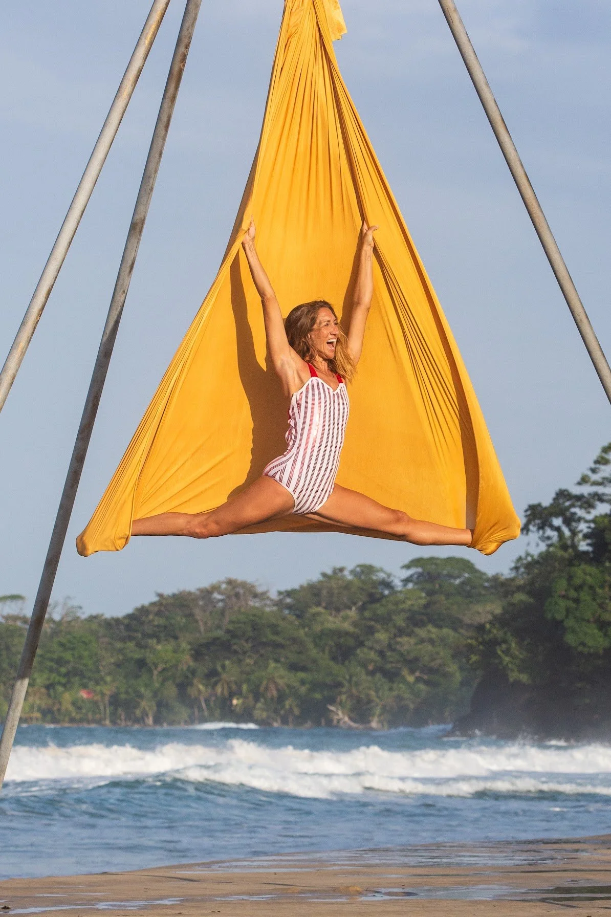 A woman doing a split yoga pose inside a yellow trapeze or hanging fabric at the beach with trees and waves in the background.