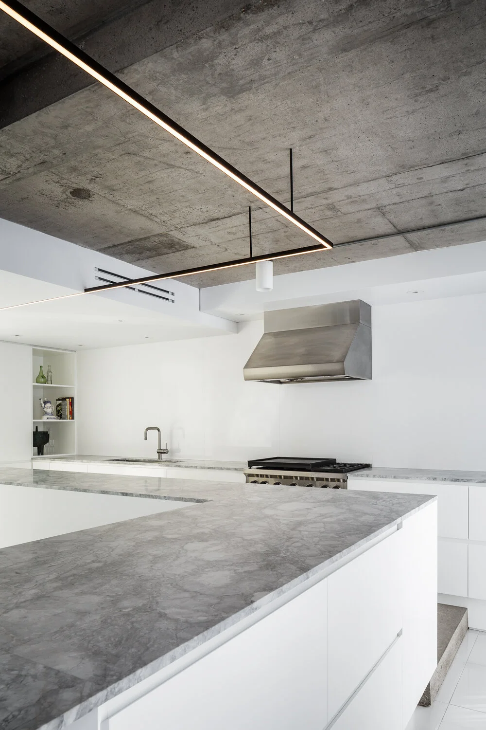 L-shaped kitchen with very large statuario marble countertop. Satin white lacquered kitchen cabinet with integrated handle. A linear LED fixture following the counter hangs from the concrete ceiling