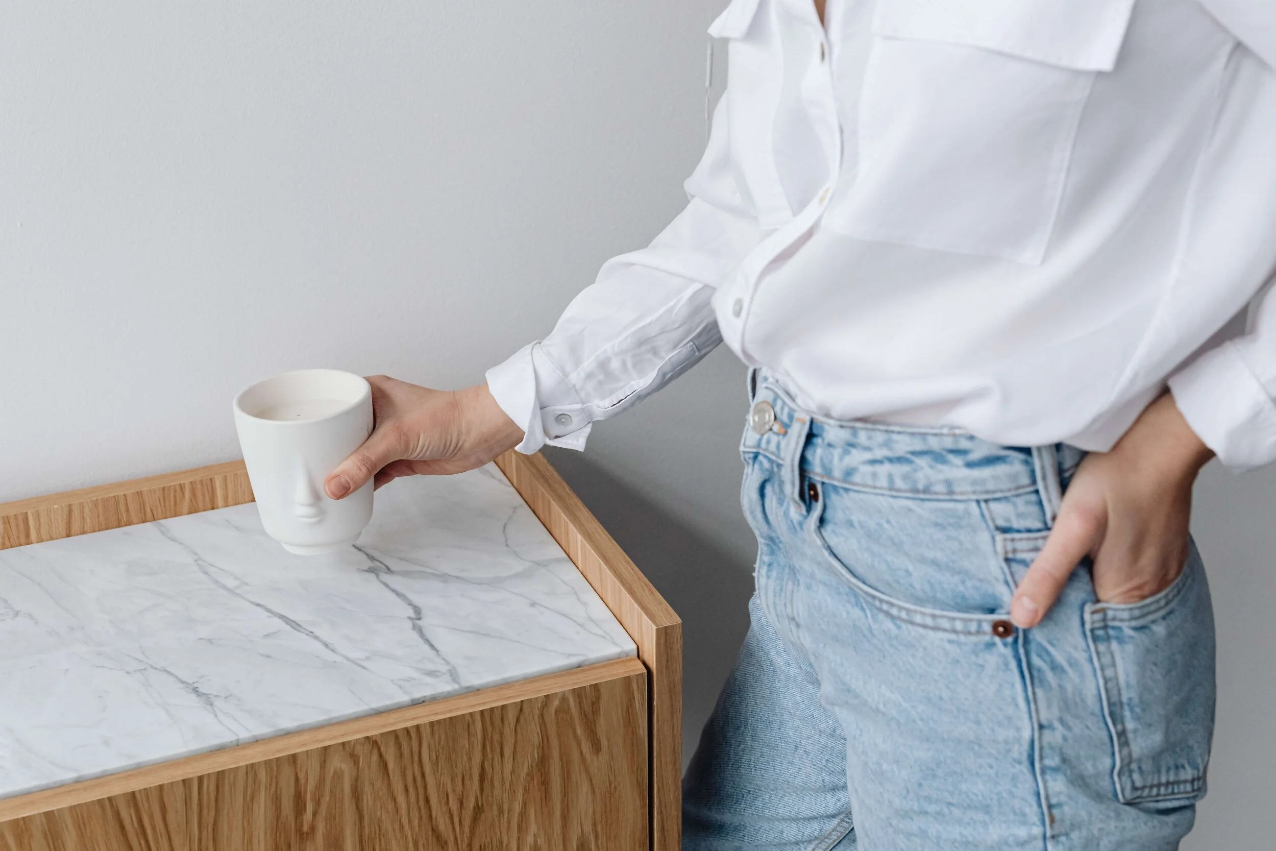 Person in white shirt and jeans holding a cup with a face design standing next to a wooden table with a marble top.