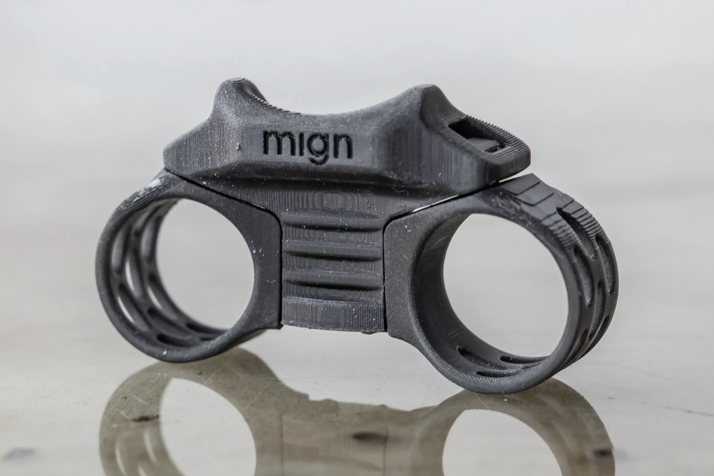 A black, 3D-printed dual-chambered object with the word 'mign' on top, featuring textured grips and a futuristic, mechanical design, placed on a reflective surface.