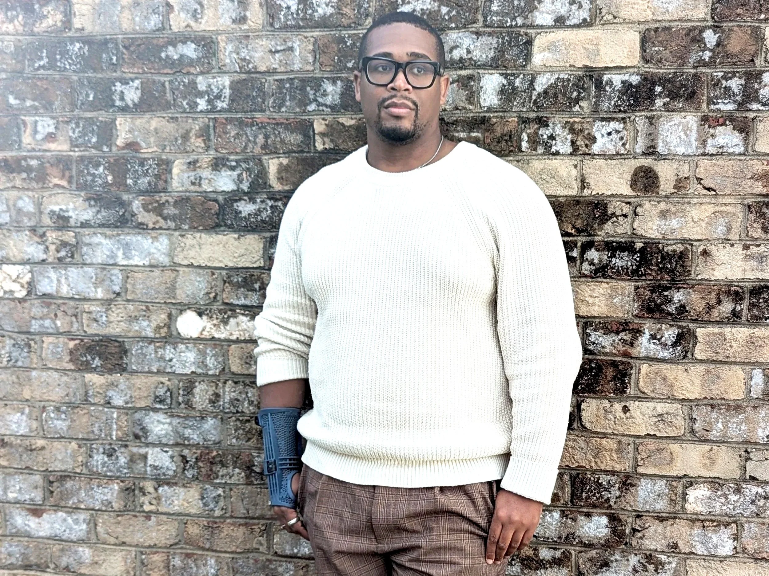 A man wearing glasses, a white sweater, and plaid pants standing against a brick wall, wearing a mign upper extremity brace on his right hand.