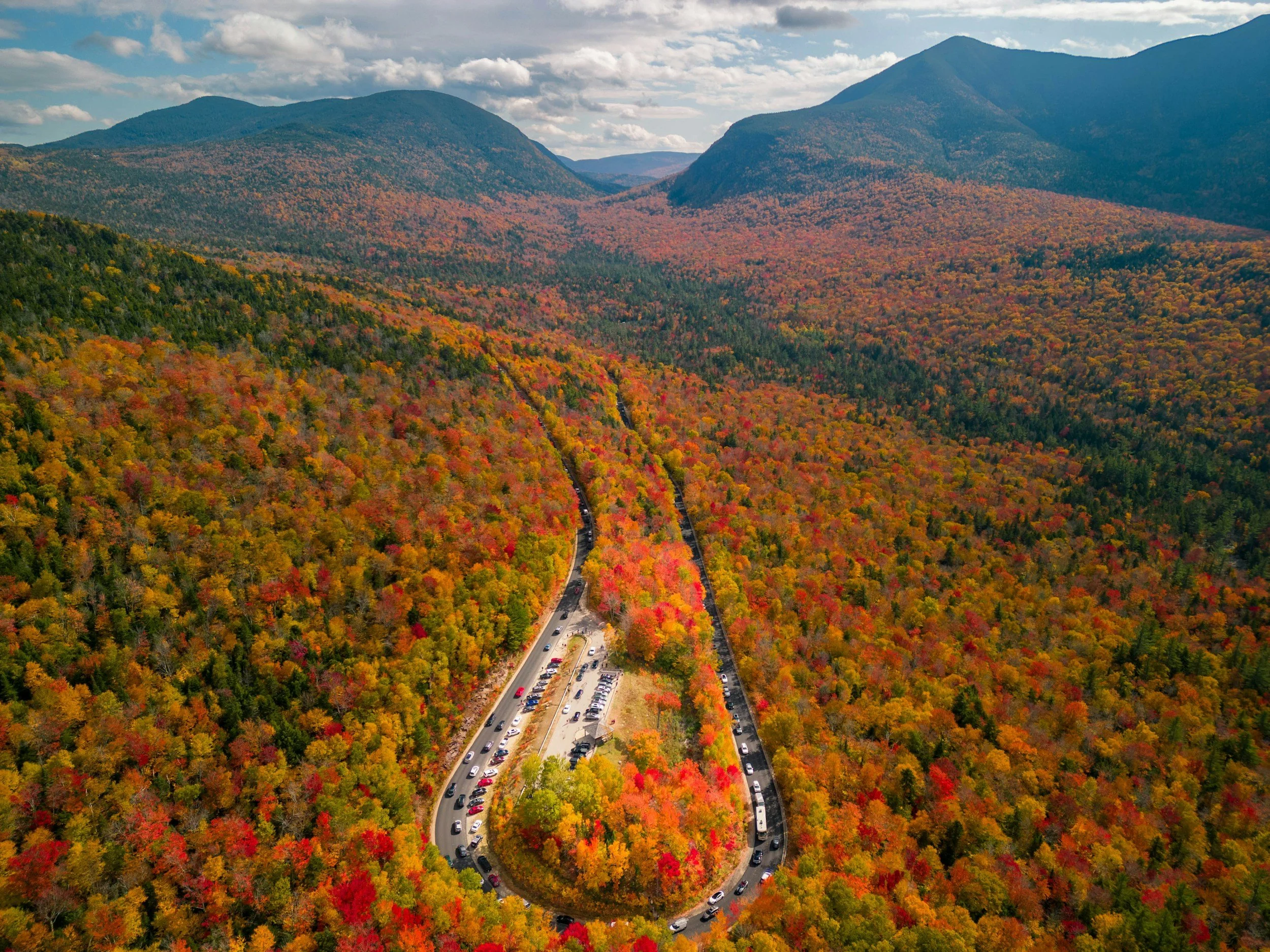 Aerial view of a winding road through a forest with colorful autumn leaves, mountains in the background, and a cloudy sky.