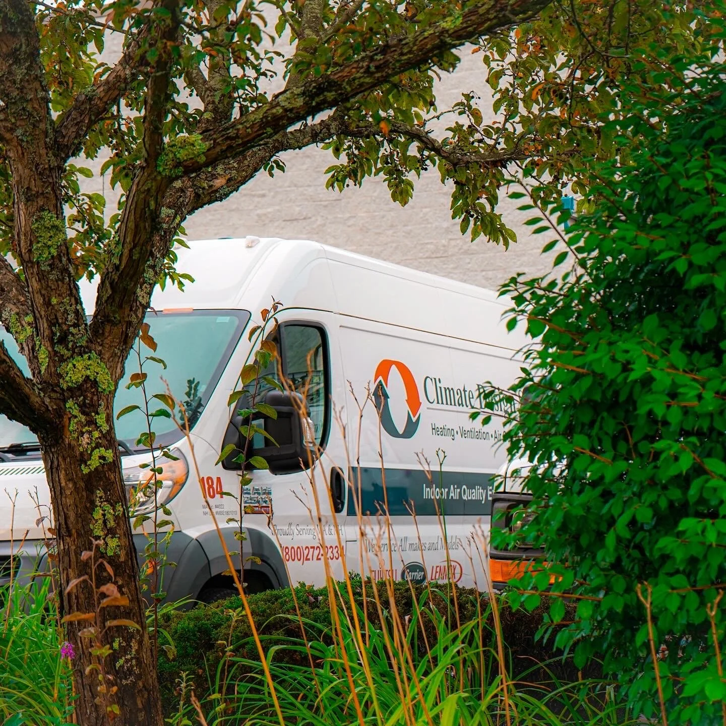 A white service vehicle with an orange and blue logo and the text 'Climate' partially visible on the side, parked behind green bushes and trees.