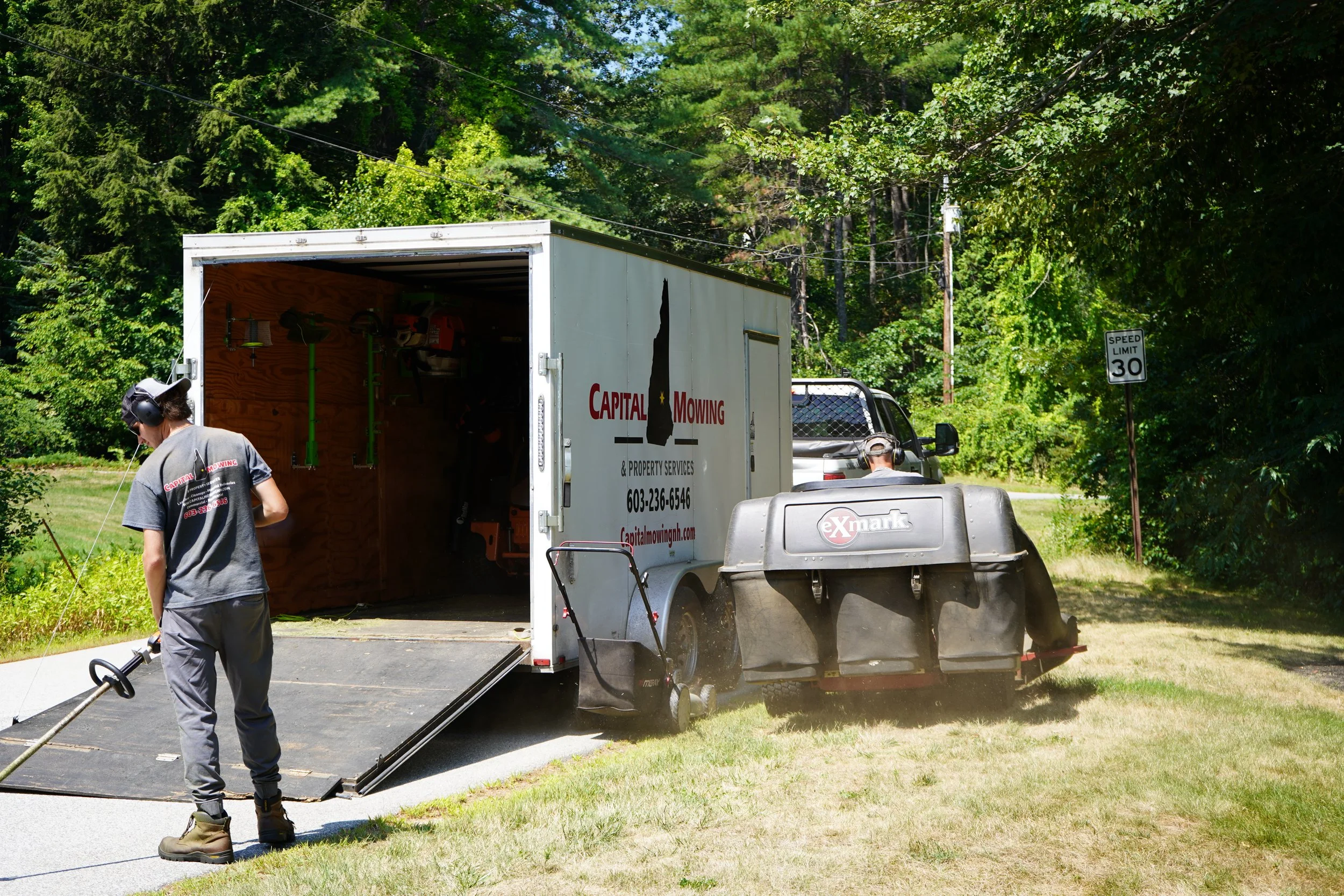 A white moving truck with open back doors parked on a driveway surrounded by green trees. A person wearing headphones, a gray t-shirt, and gray pants is using a grass trimmer. The truck has the words "Capital Mowing & Property Services" on its side.