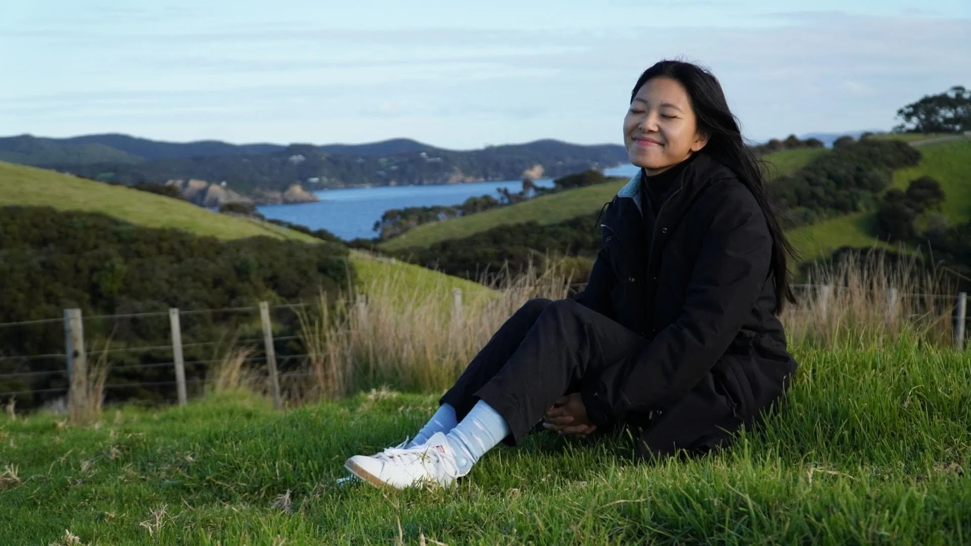 Young woman sitting on grassy hill with eyes closed and a gentle smile, overlooking green rolling hills, trees, and a body of water in the distance on a partly cloudy day.