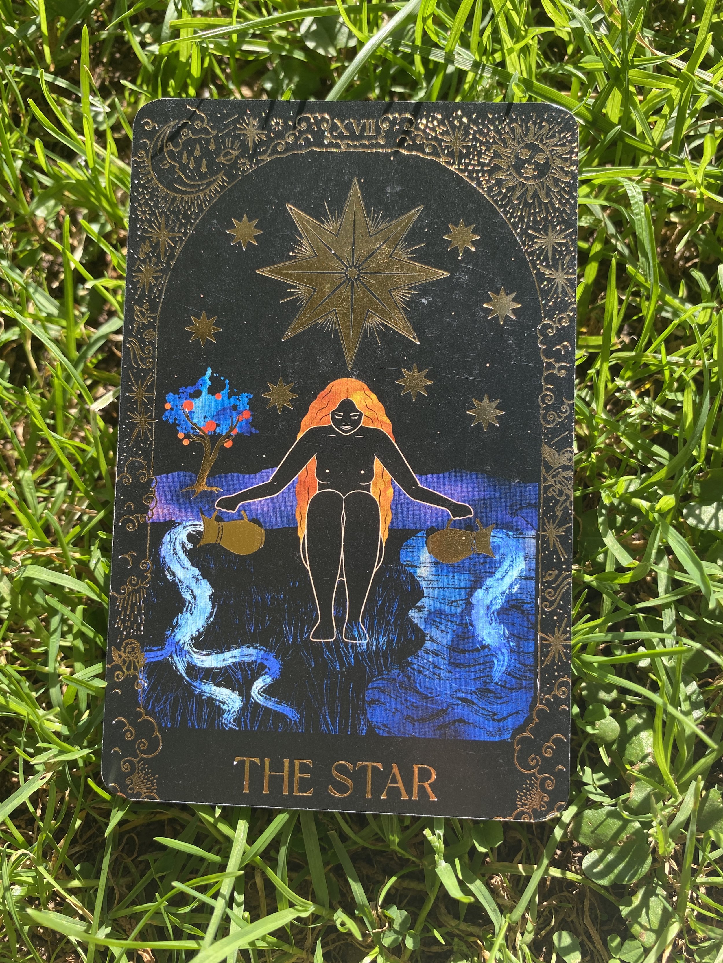 Tarot card titled 'The Star' depicting a woman with orange hair floating above water, holding two watering cans, with a large star and smaller stars above her, a tree with red fruit to her side, and a river flowing in front of her.