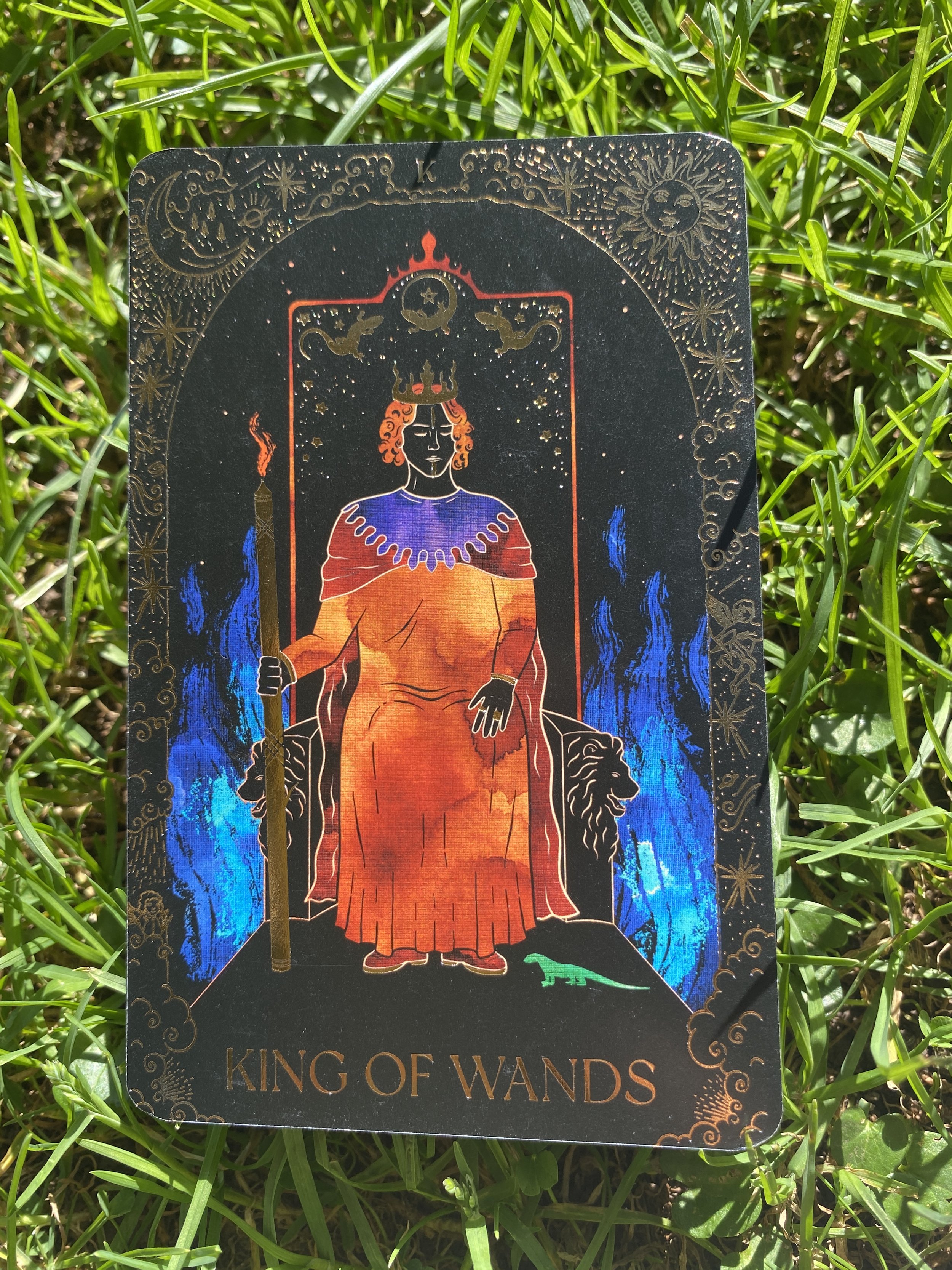 The tarot card titled 'King of Wands' features a regal figure seated on a throne, holding a staff, with a crown on his head, surrounded by flames and symbols of power, with a lion on one side and a small lizard at his feet. The background is dark with celestial imagery.