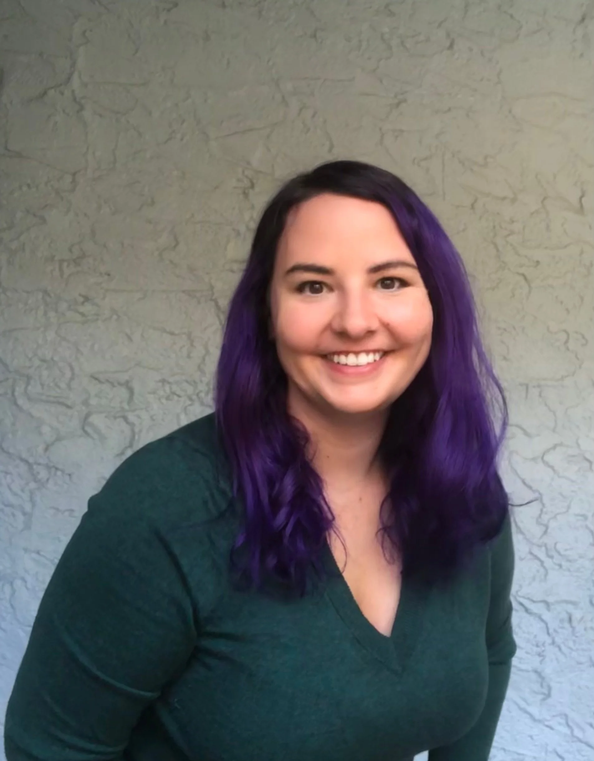 A woman with purple hair smiling, wearing a dark green top, in front of a textured light gray wall.