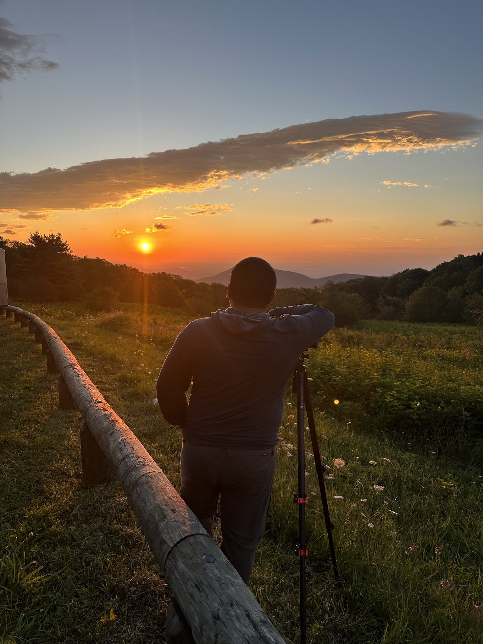 Person standing on a grassy hill, looking at a sunset over distant mountains, with a camera on a tripod, and a wooden fence along the path.