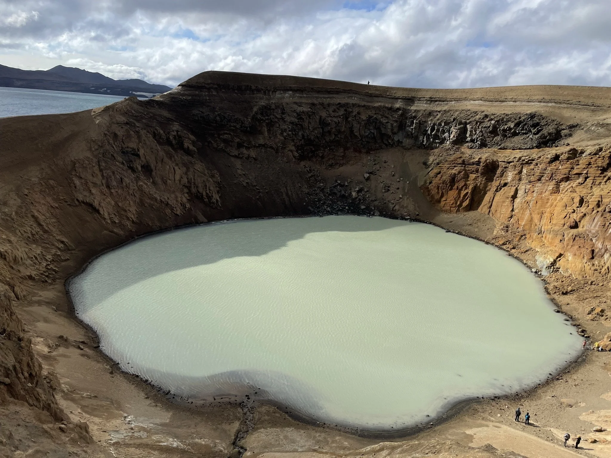 A large crater lake with milky turquoise water, surrounded by rugged, reddish-brown rocky terrain and cliffs, with a few people near the edge for scale.