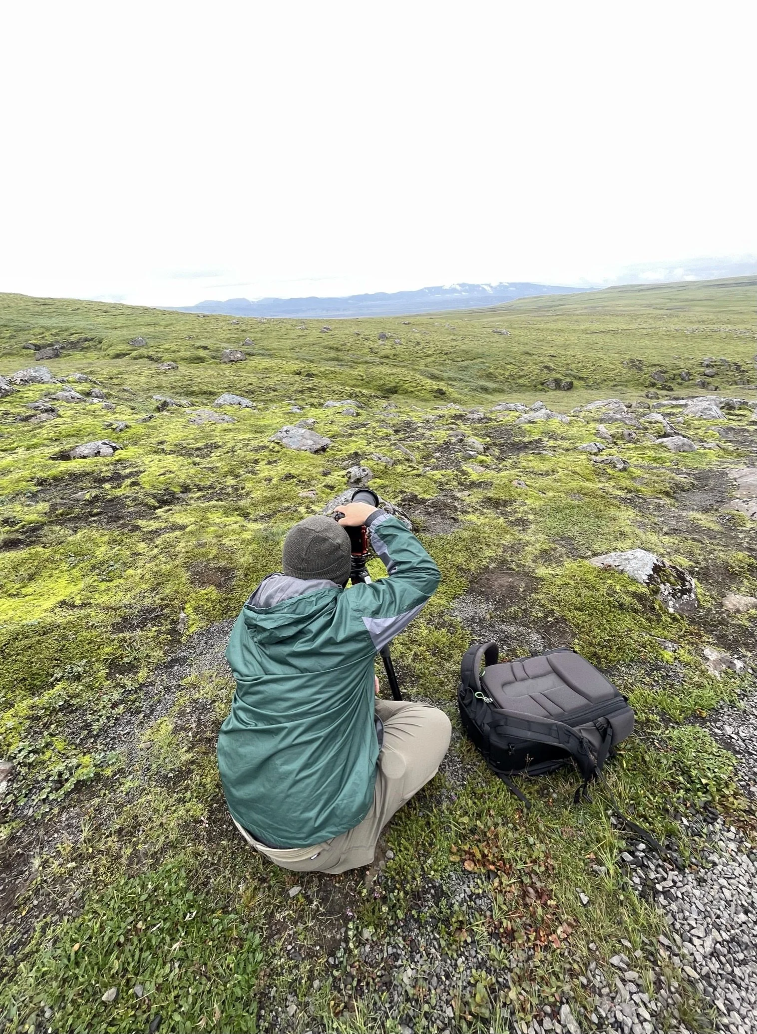 Person in green jacket and beige pants kneeling on mossy ground, looking through binoculars, with a camera bag nearby, in a vast green landscape under an overcast sky.