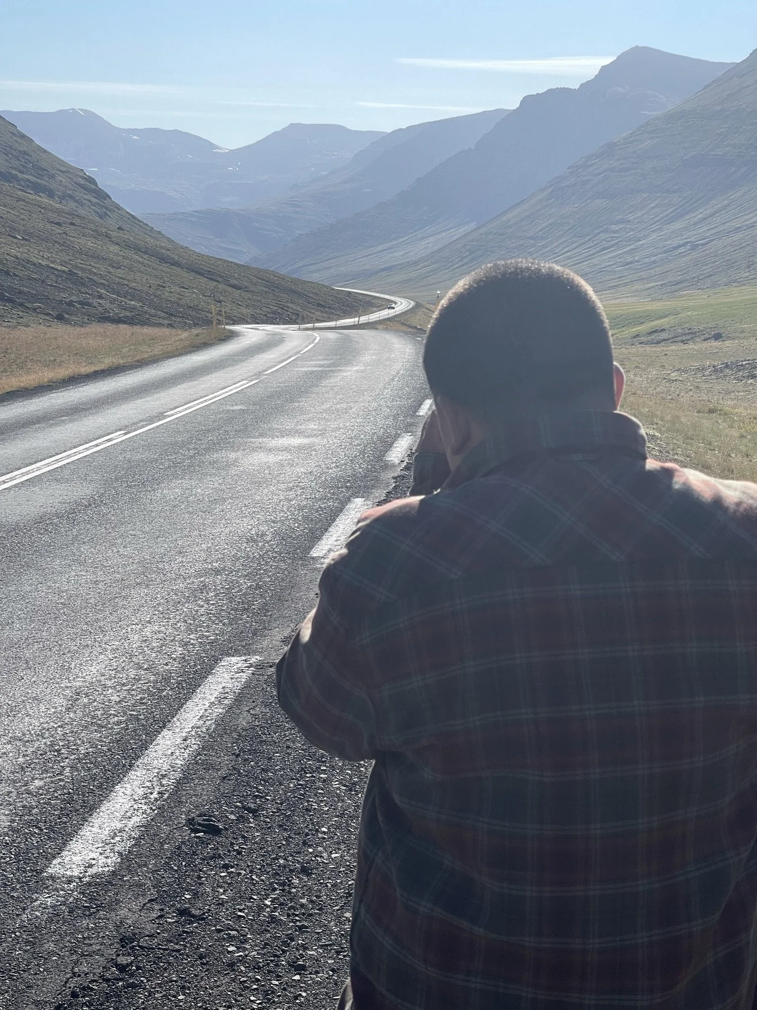 A person in a plaid shirt taking a photo or looking down at a long, winding mountain road with mountains in the background.
