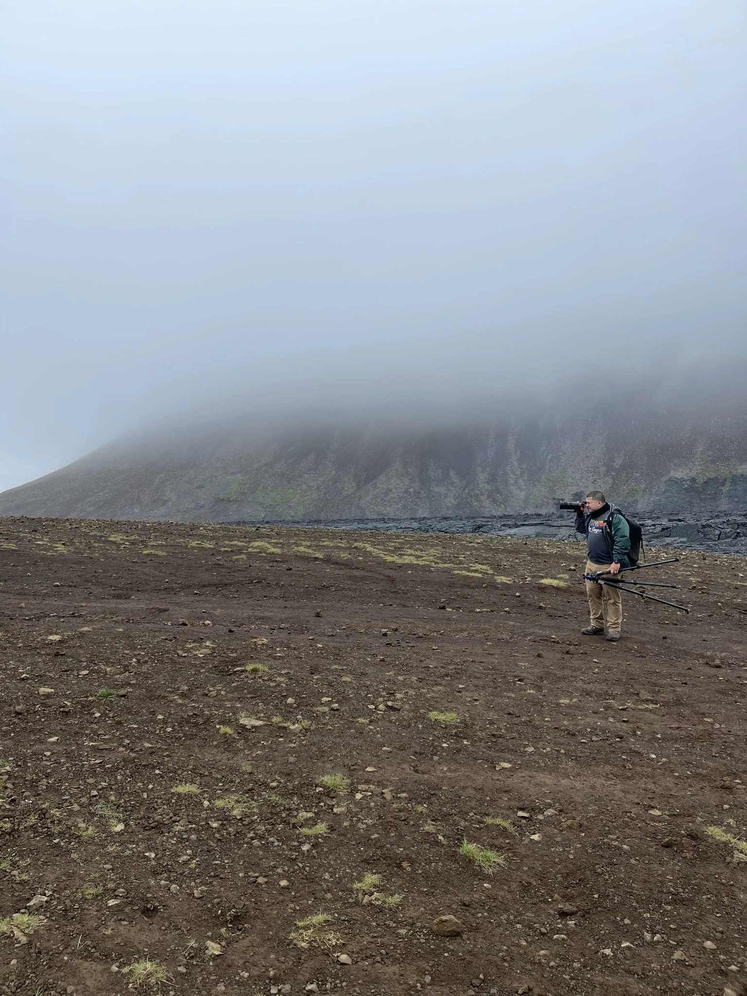 A person with a camera and tripod standing on a barren, rocky landscape with sparse grass, in front of a foggy mountain.