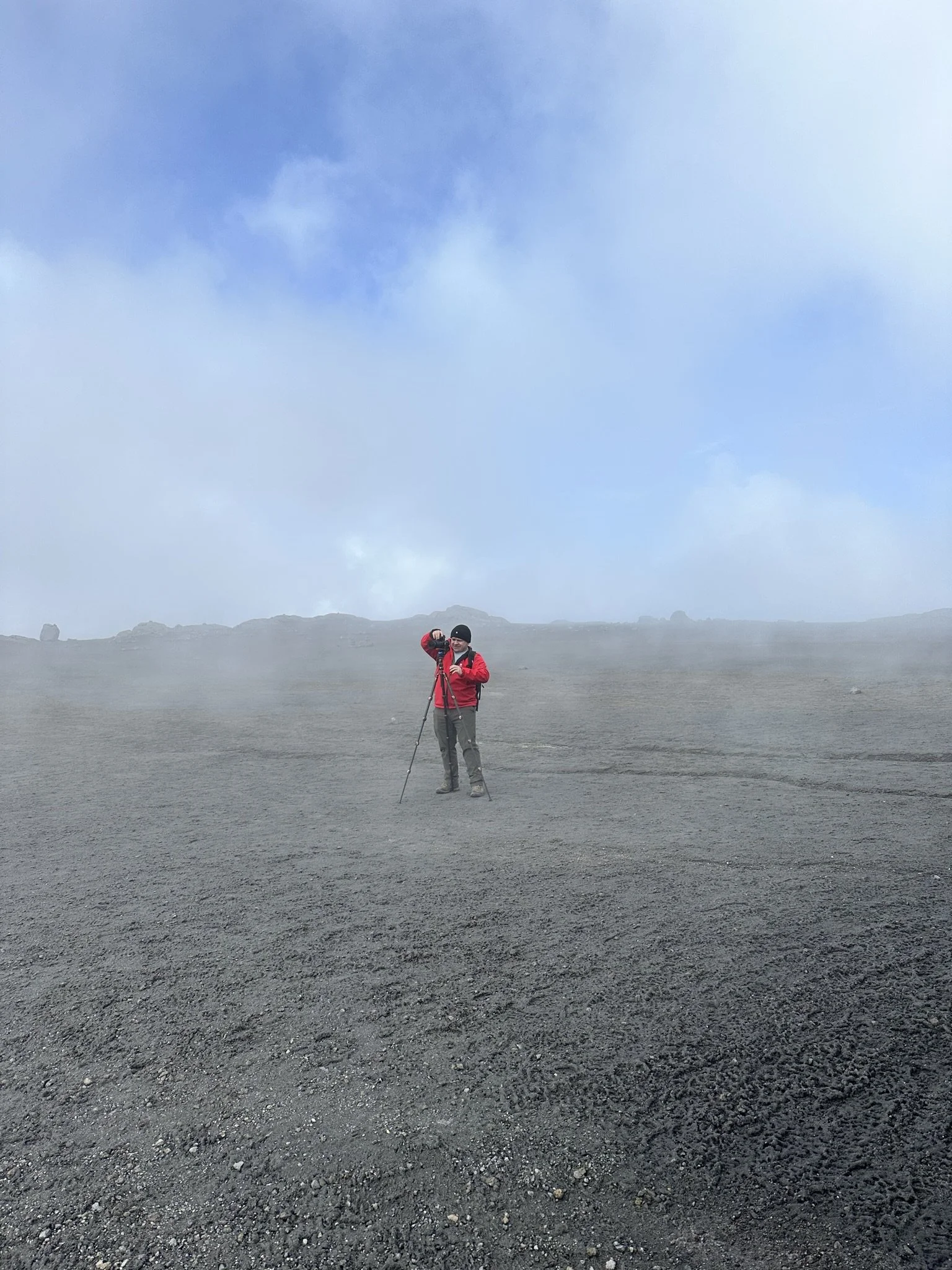 A person dressed in outdoor gear, including a red jacket, standing on dark volcanic or gravelly terrain, using hiking poles in a foggy, open landscape with blue sky and some clouds overhead.