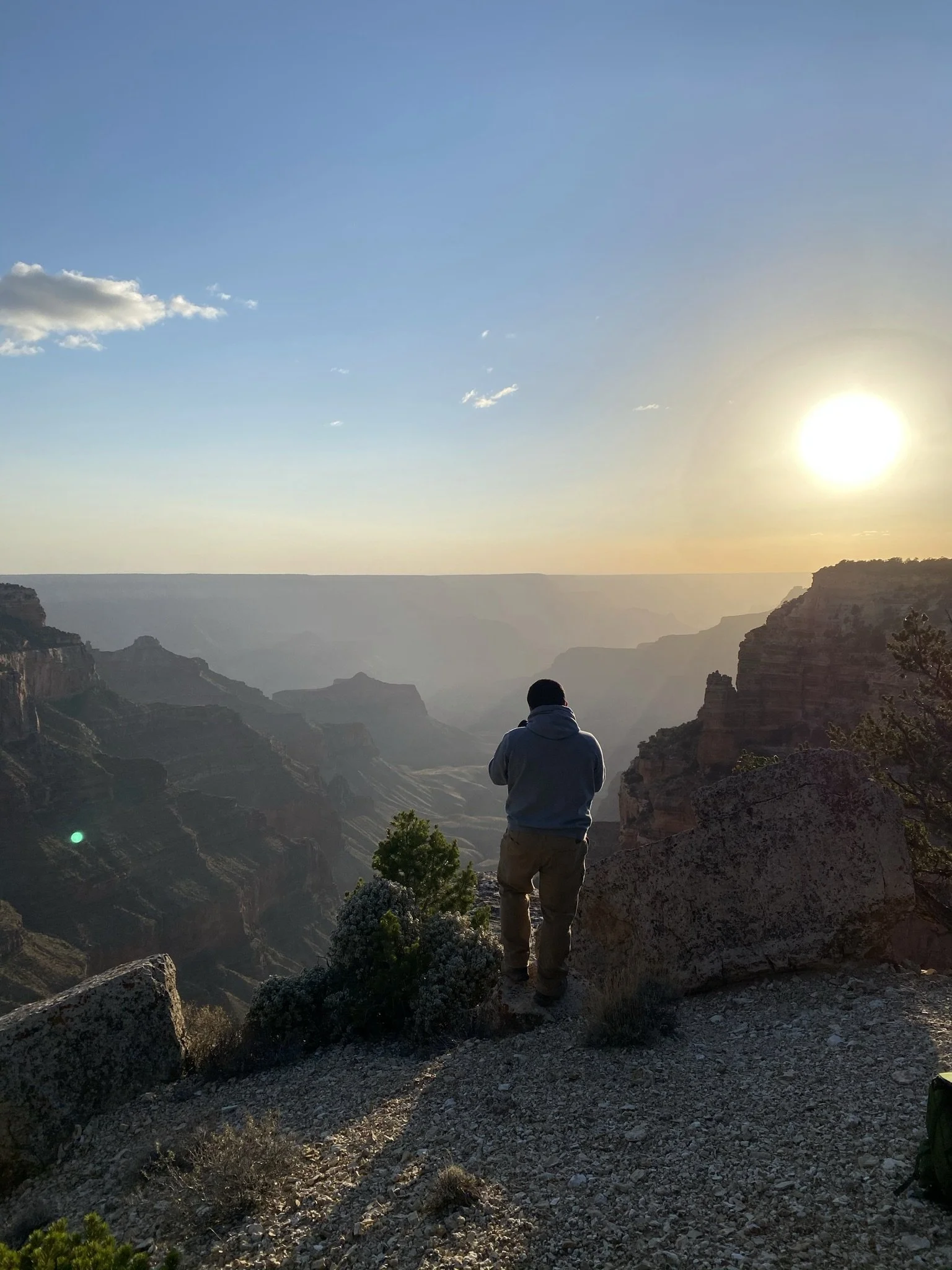 A person standing on rocky terrain overlooking the Grand Canyon at sunset, with the sun low in the sky and a few clouds.