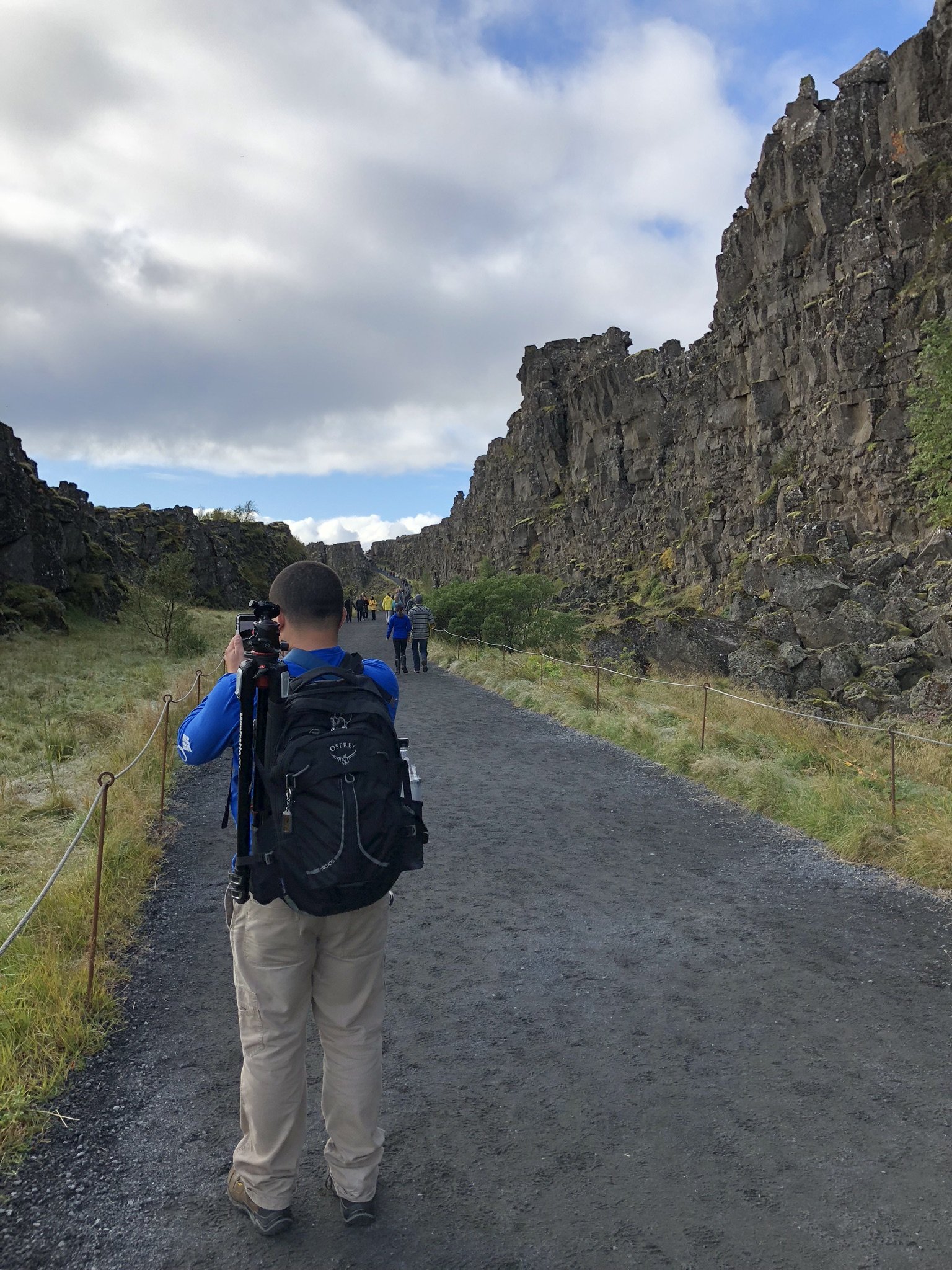 Person with a backpack taking a photo on a gravel path between rocky cliffs surrounded by green grass and small trees, with a cloudy sky overhead.