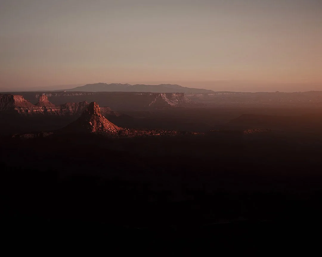 Sunset over a rocky canyon landscape with mesas and plateaus in the distance.