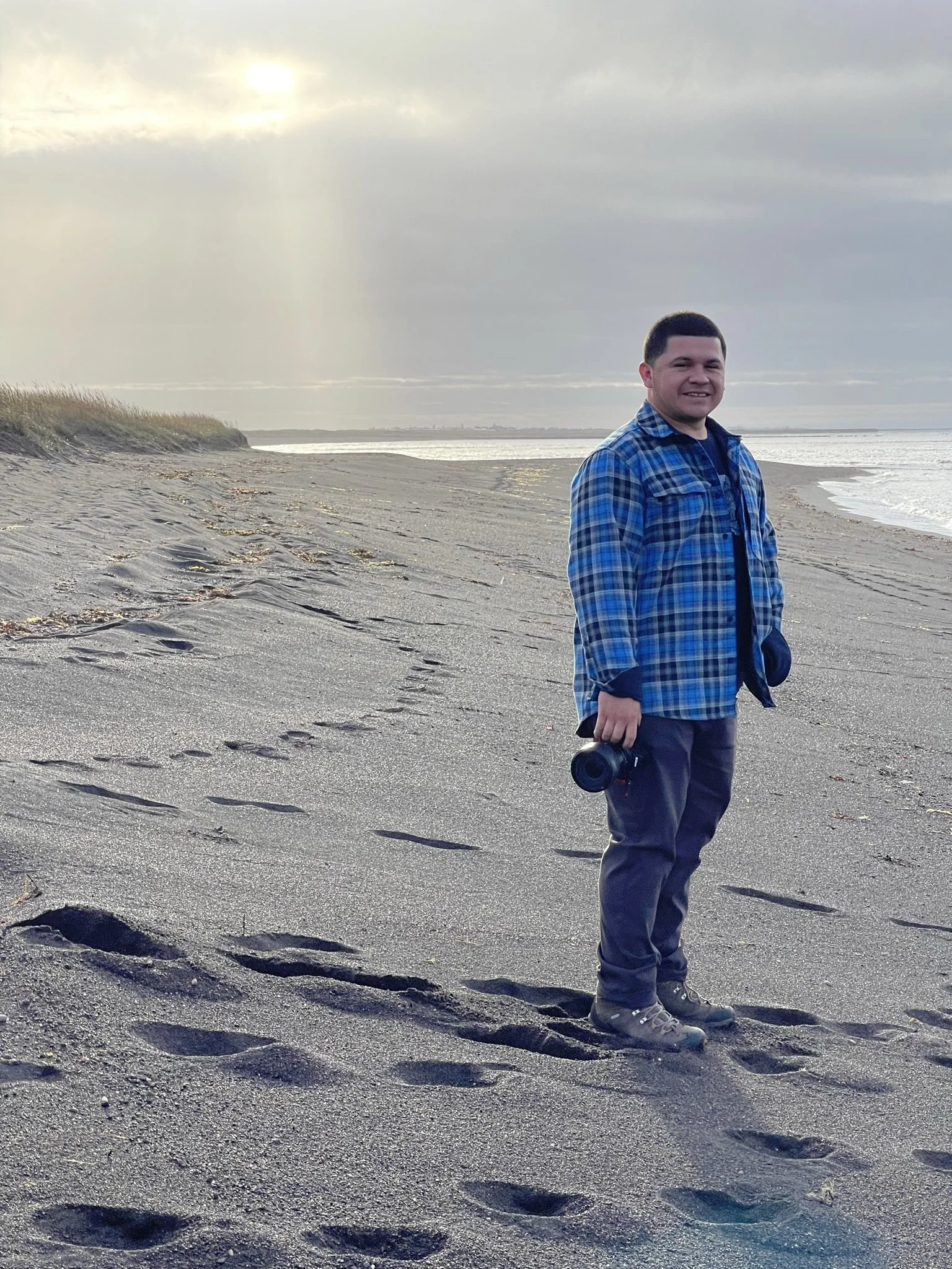 A man standing on a sandy beach, smiling, holding a camera, wearing a blue plaid shirt, dark pants, and hiking boots, with footprints in the sand and the ocean in the background under a cloudy sky.
