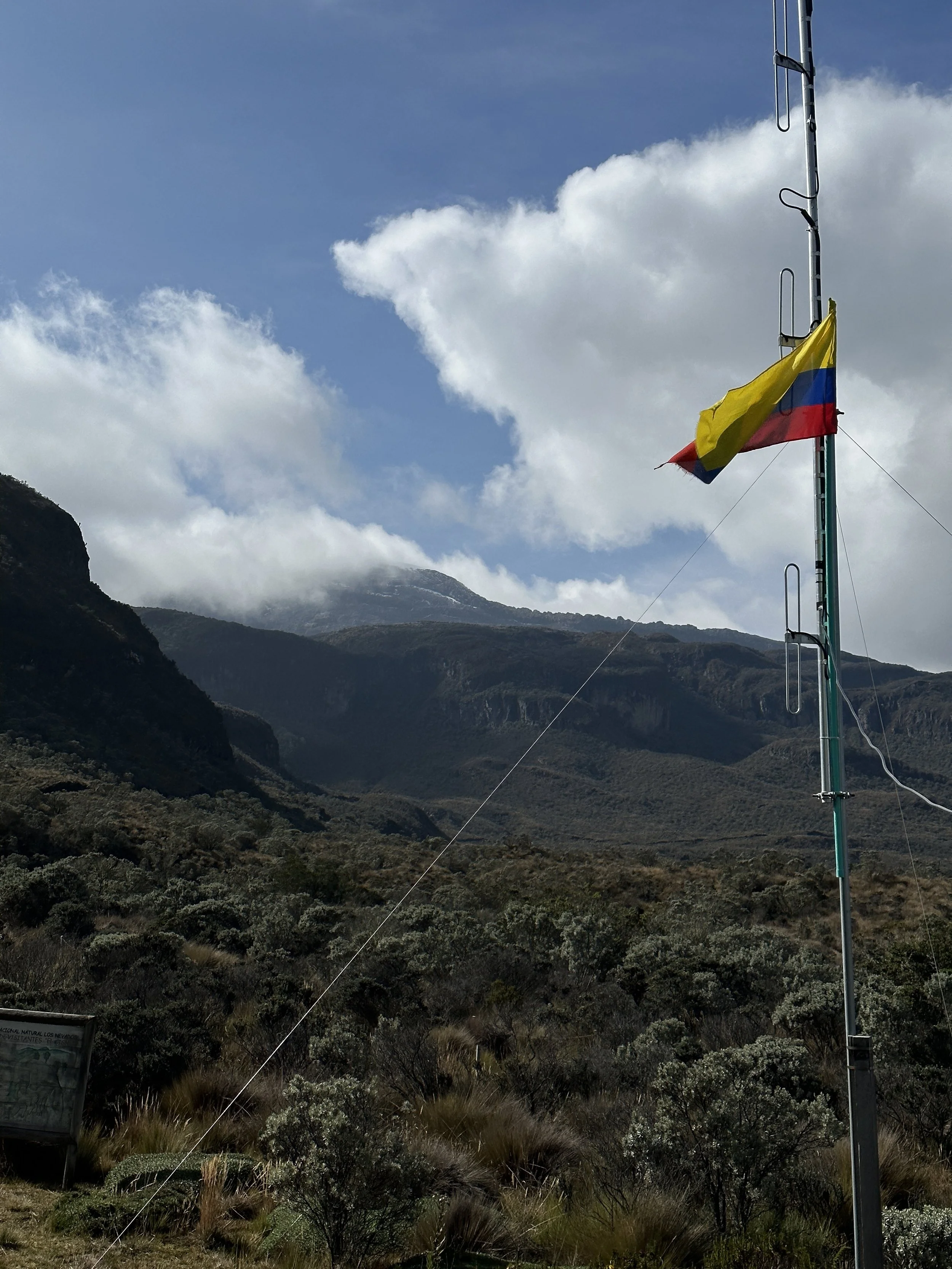 Flagpole with the Colombian flag in a mountainous landscape with clouds and forested terrain.