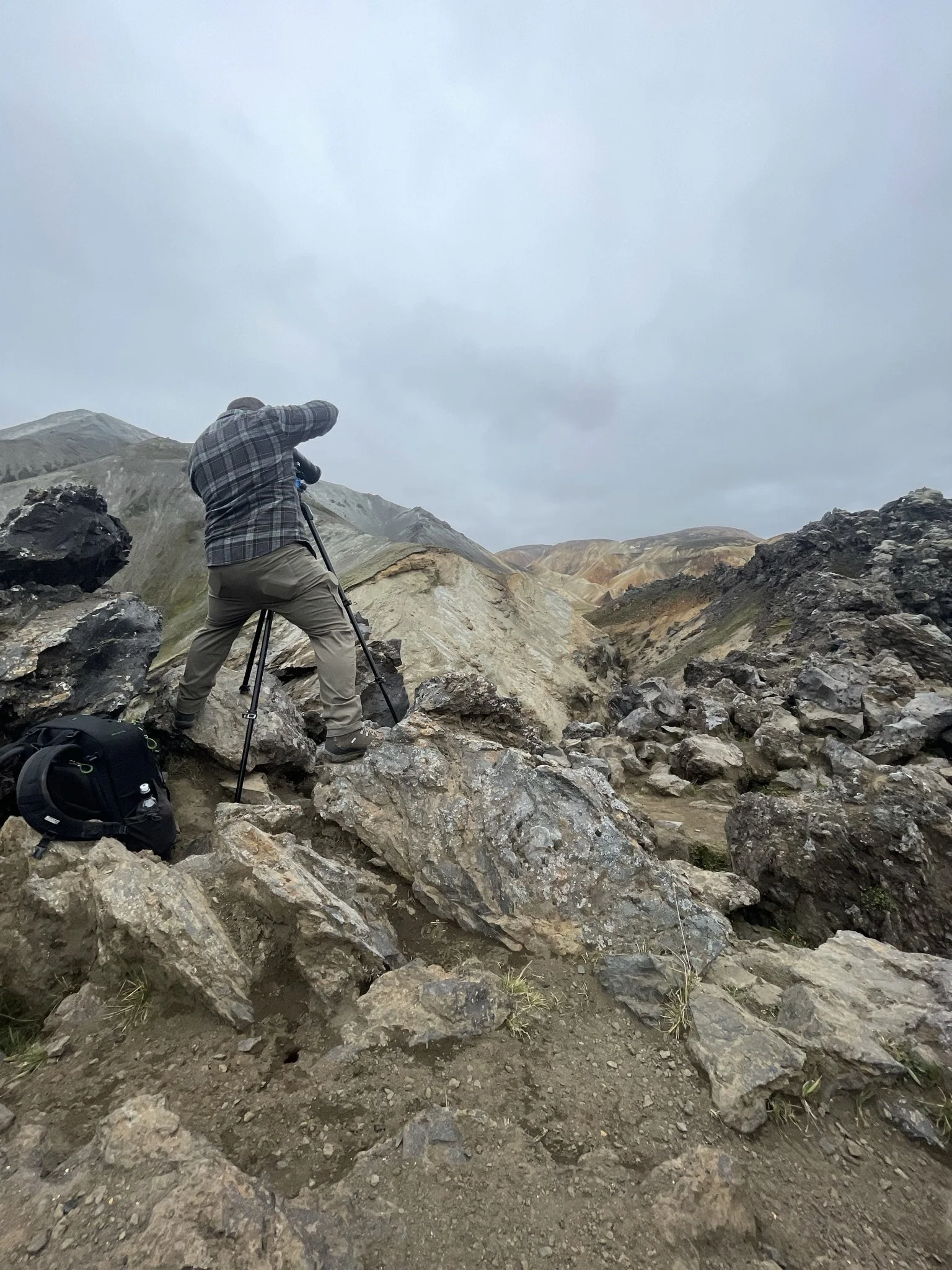 A person with a camera tripod taking photos on rocky, mountainous terrain under a cloudy sky.