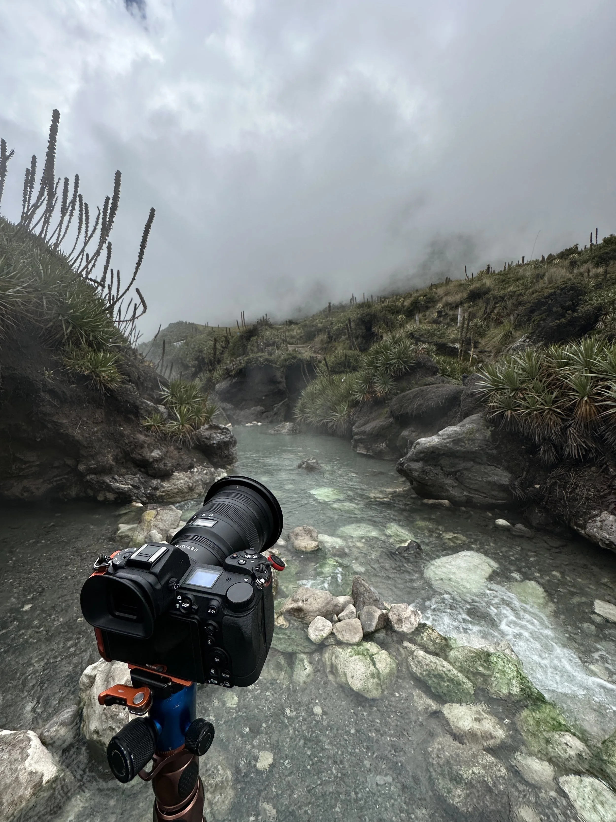 A camera mounted on a tripod overlooking a hot spring with rocky edges, surrounded by green vegetation and cacti, under a cloudy, foggy sky in a mountainous area.