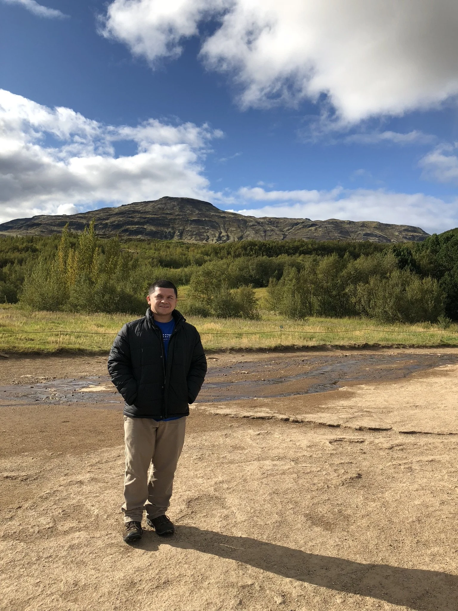 A man standing outdoors on a dirt surface with his hands in his pockets, wearing a black jacket and beige pants. Behind him, there are green trees and a mountain under a partly cloudy sky.