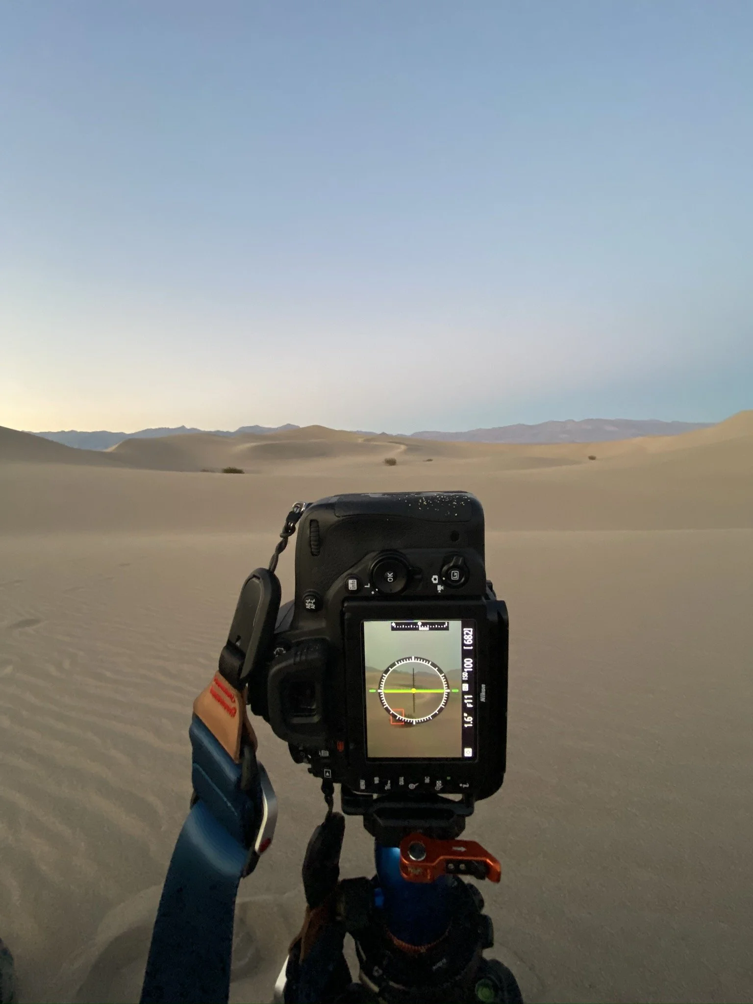 A digital camera mounted on a tripod captures a desert landscape with sand dunes and mountains in the background during sunset or sunrise.