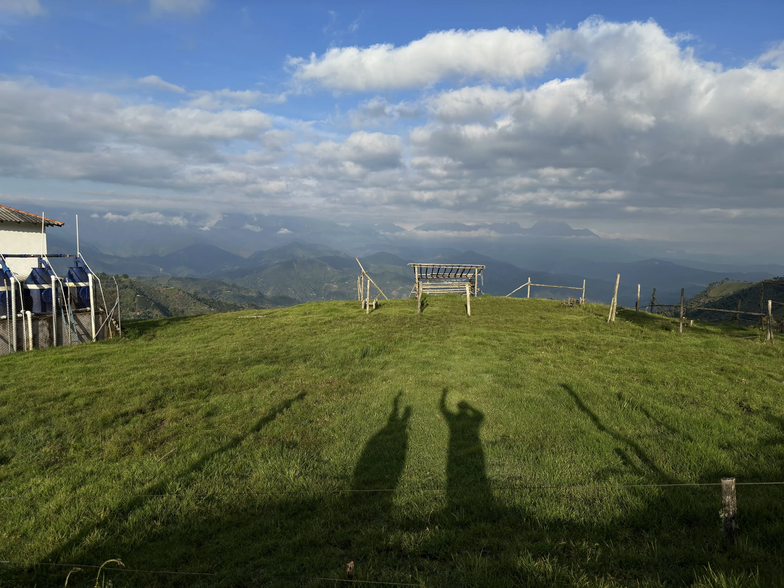 A scenic hillside with a grassy area, a bench, and mountain views in the distance. Two shadows of people can be seen on the grass, with a partly cloudy sky overhead.