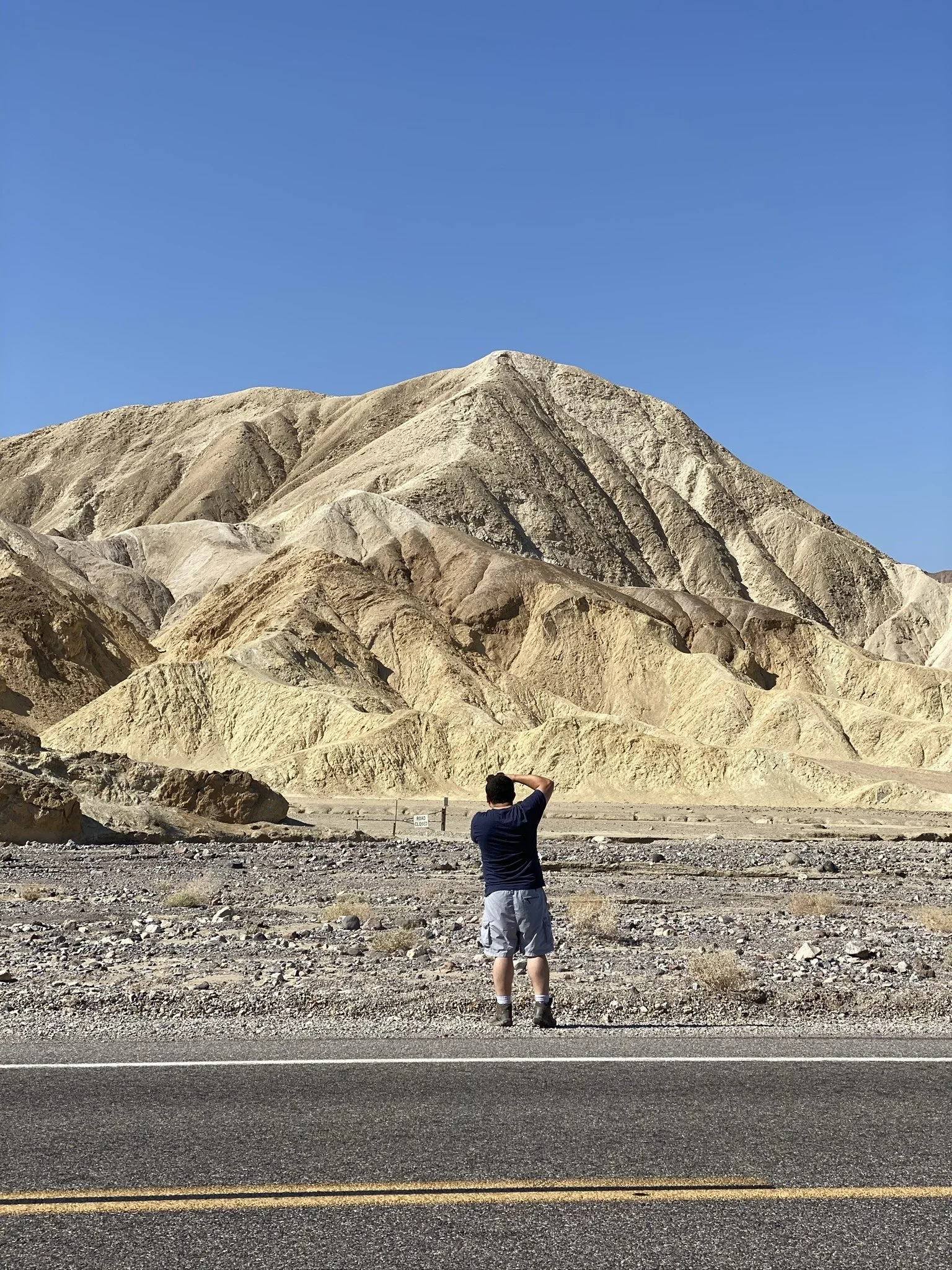 A man wearing a navy shirt, shorts, and hiking boots standing on a roadside, looking at a large mountainous desert landscape with arid, rocky hills under a clear blue sky.