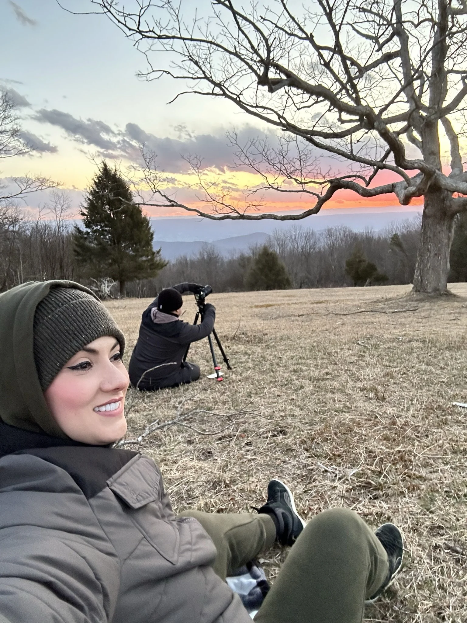 Two people sitting on the ground in a field during sunset, one smiling at the camera and the other looking through a telescope, with large trees and a colorful sky in the background.