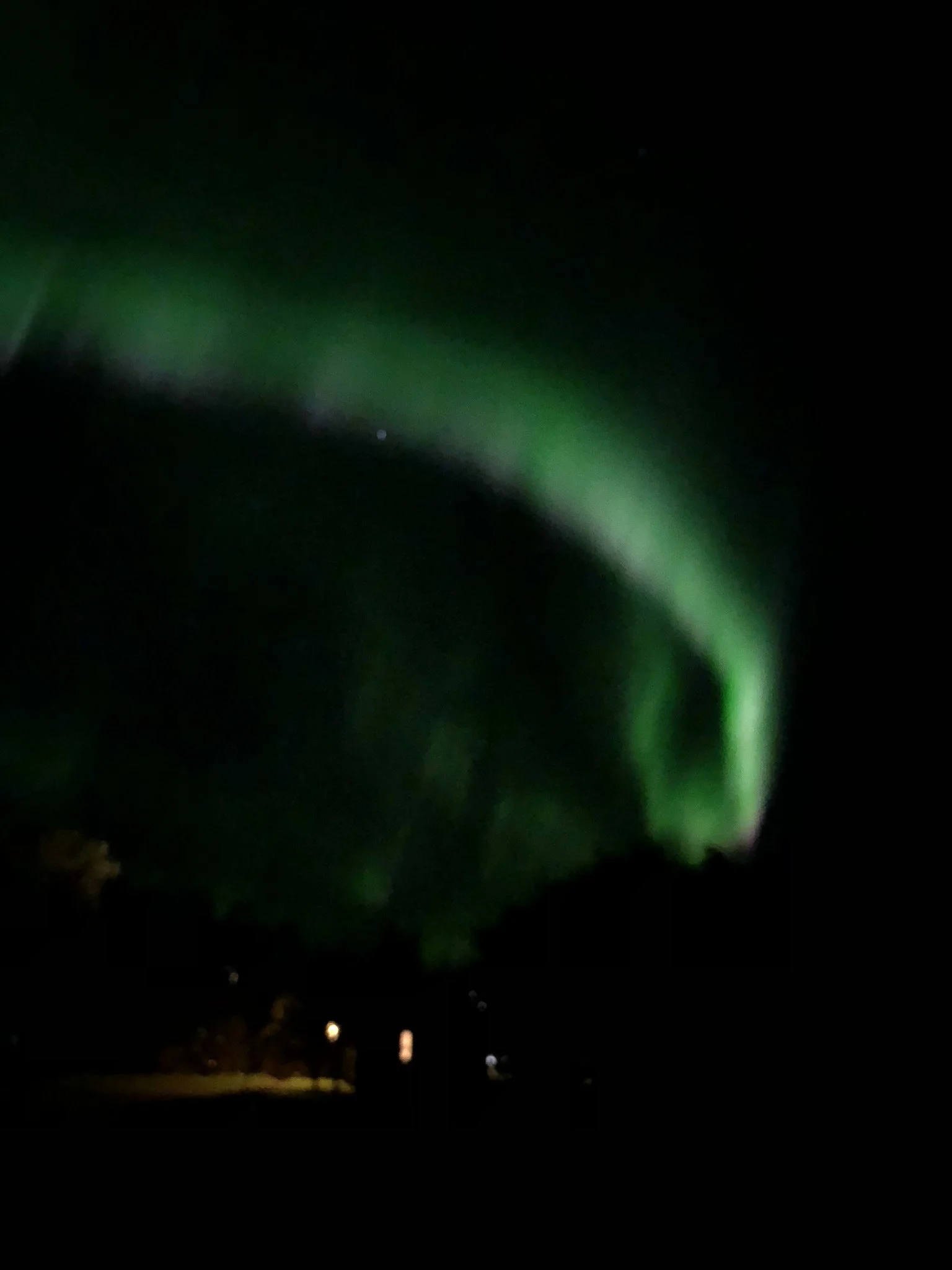 Northern lights illuminating the night sky in green hues above a dark landscape with some distant lights.