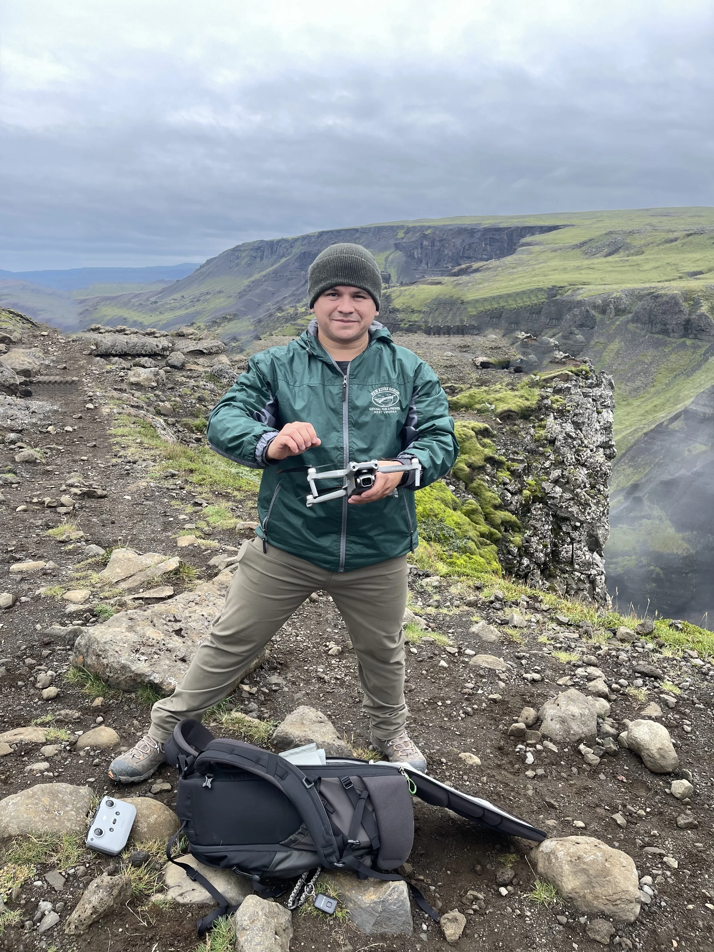 A man standing on rocky terrain with green hills and cliffs in the background, holding a drone. He is wearing a green jacket and a dark beanie. There is a black backpack and drone accessories on the ground.