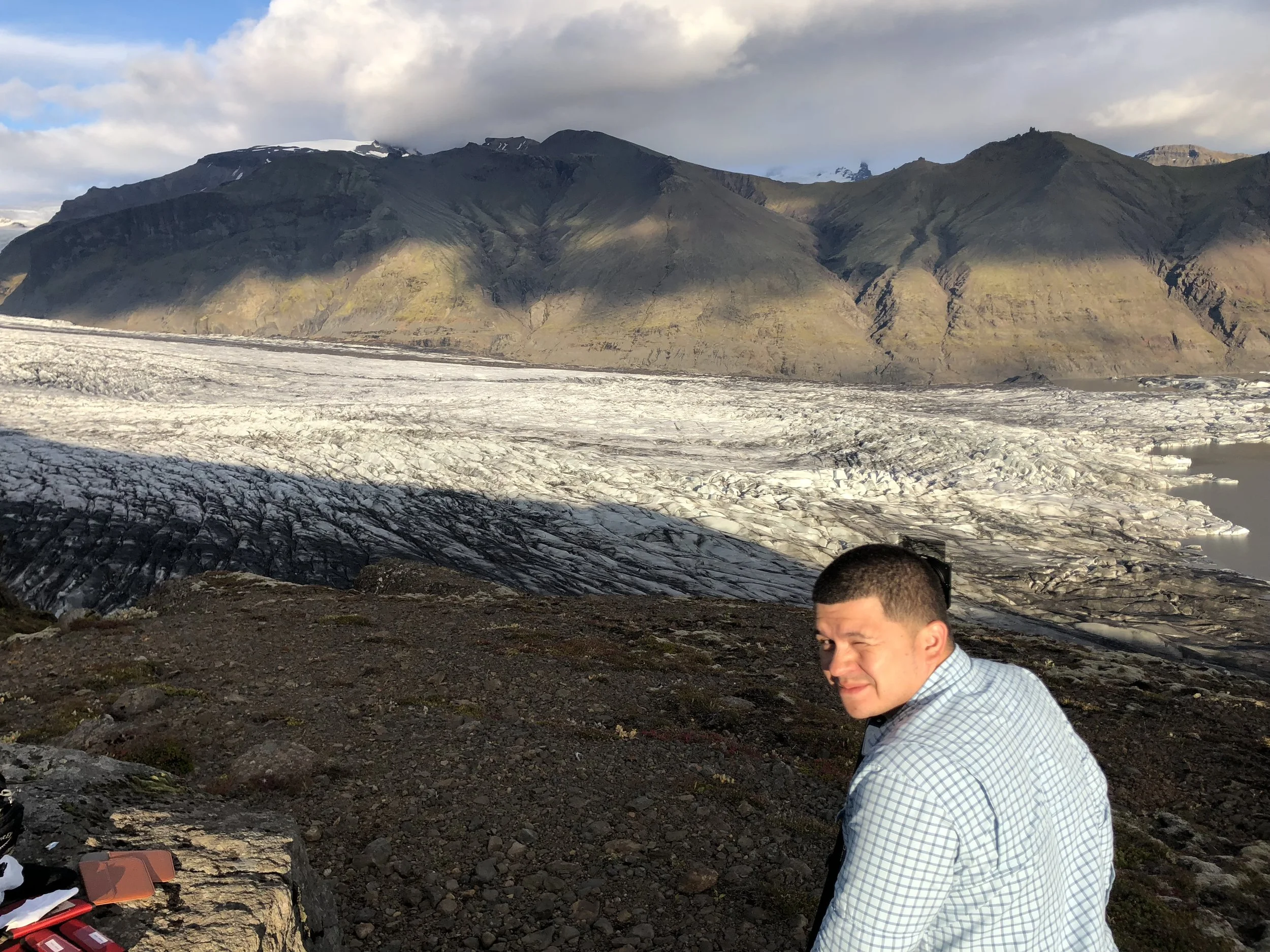 A man in a checkered shirt squinting and winking at the camera in front of a glacier with mountains in the background.