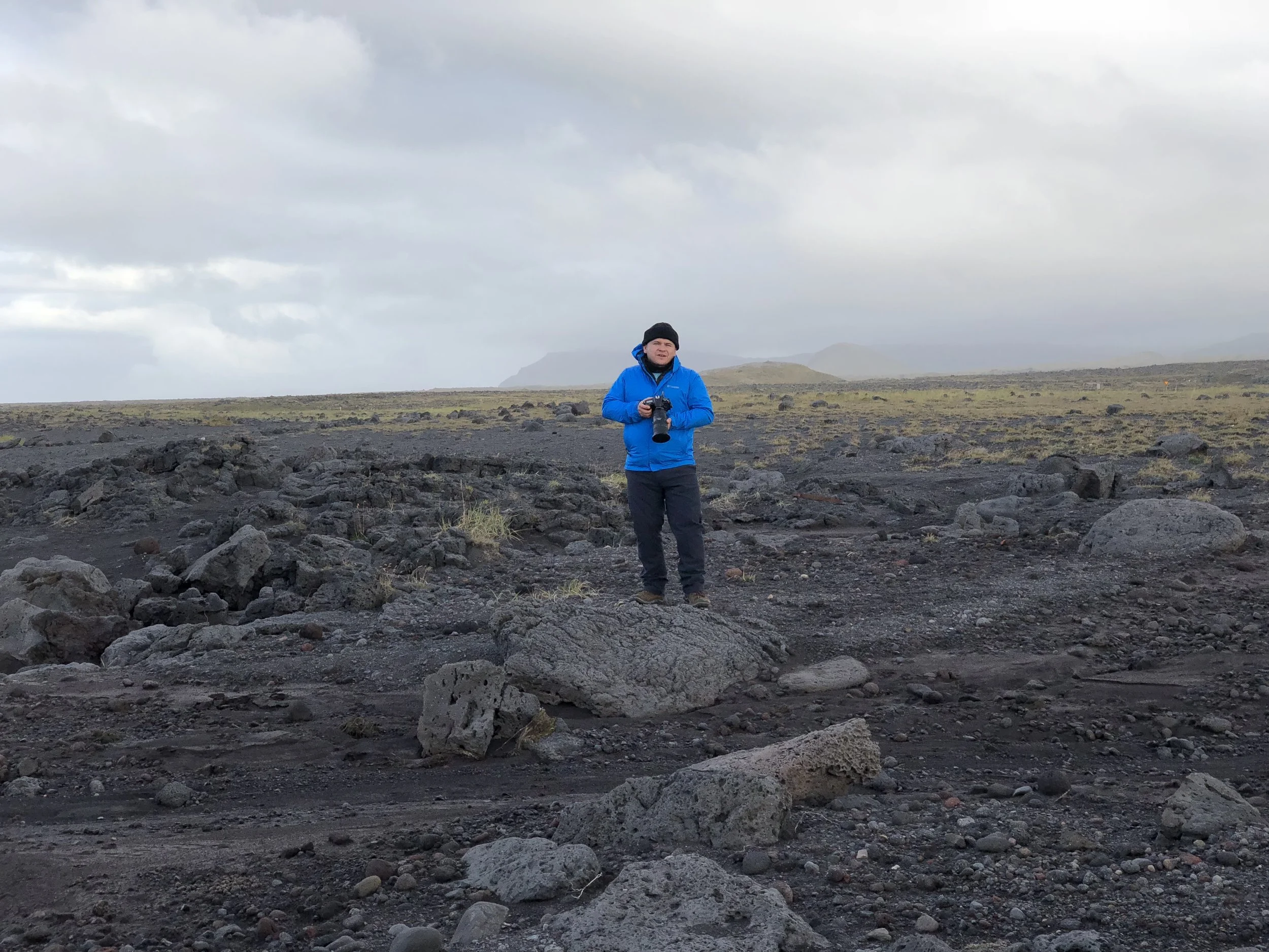 Person in a blue jacket holding a camera standing on a rocky volcanic landscape under cloudy sky.