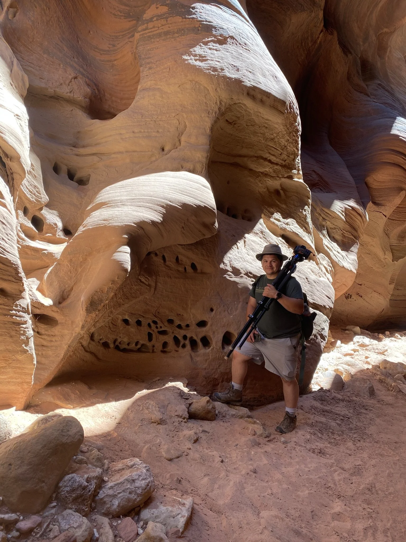 Melser Bonilla standing inside a narrow sandstone slot canyon in the Southwest USA, holding a tripod, showcasing his photography gear and adventurous spirit.