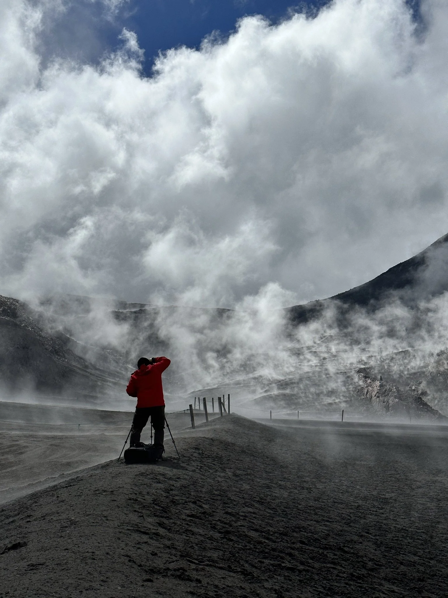 A person in a red jacket stands on a mountain trail amid clouds and mist, holding a camera or binoculars, with mountains and a cloudy sky in the background.