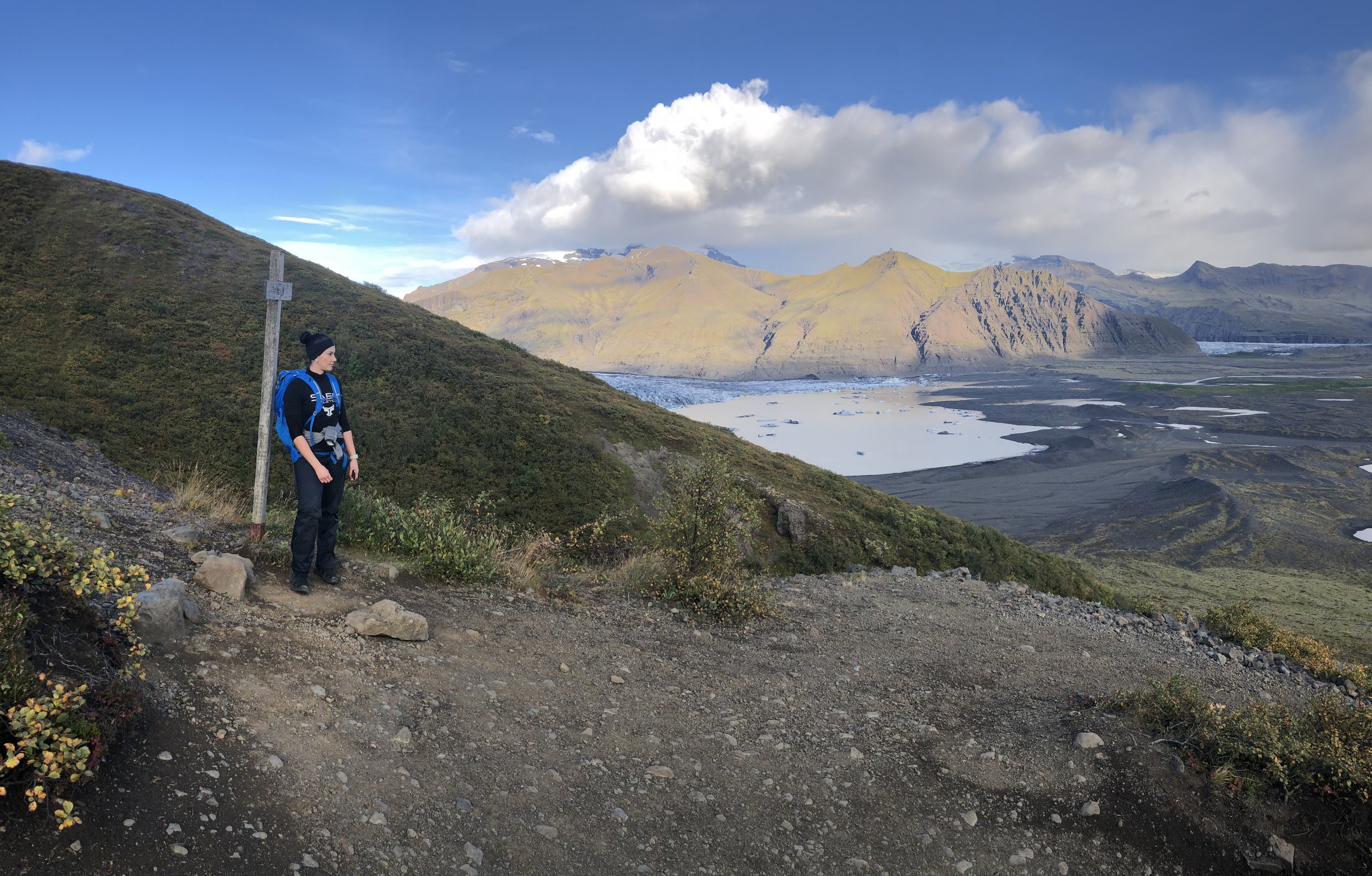 A person with a black beanie and blue backpack standing on a mountain trail overlooking a lake and mountainous landscape with partly cloudy sky.