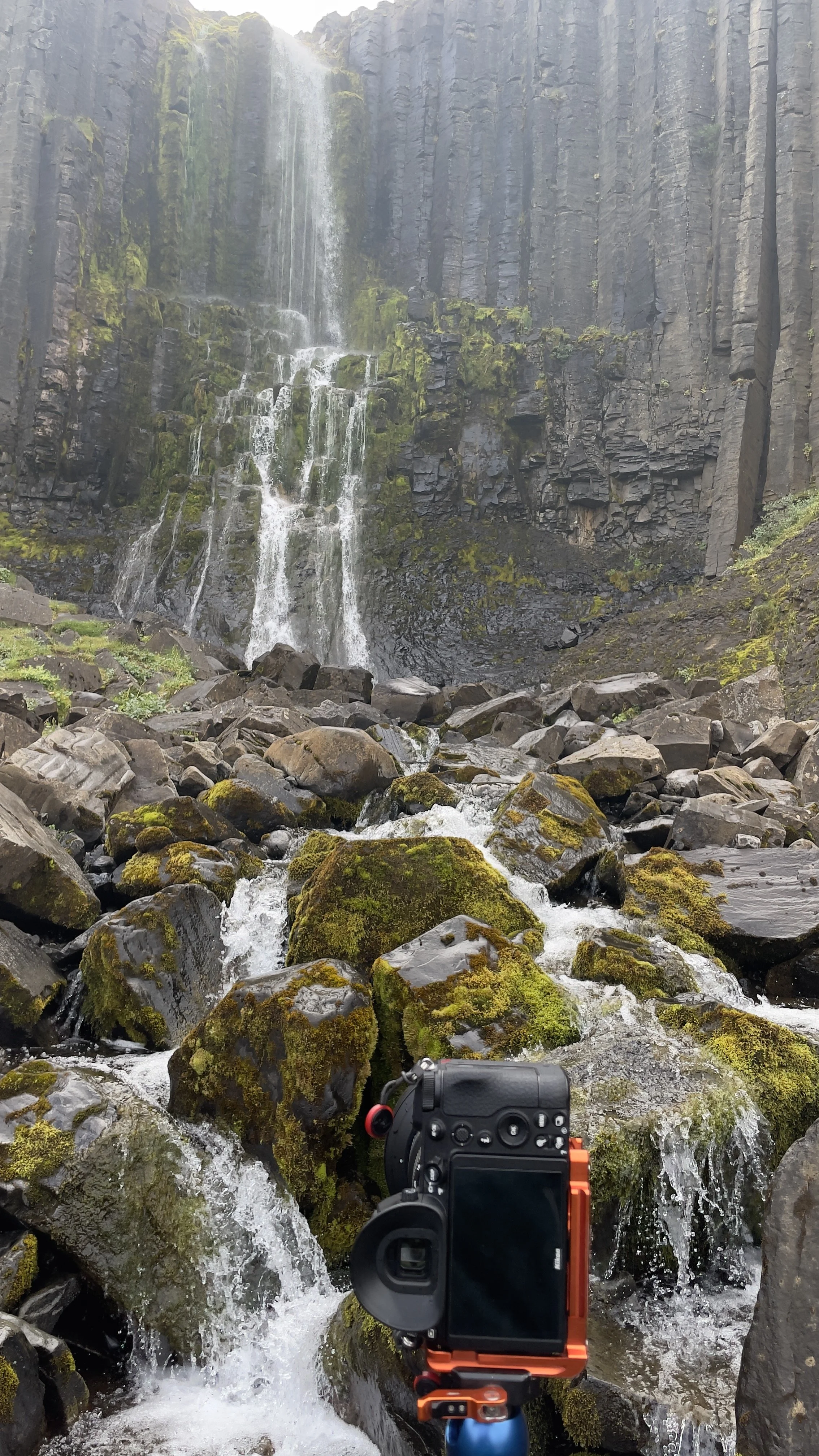 A waterfall cascading down a cliff into a rocky stream, with moss-covered stones, a camera mounted on a tripod in the foreground, in a lush, green landscape.