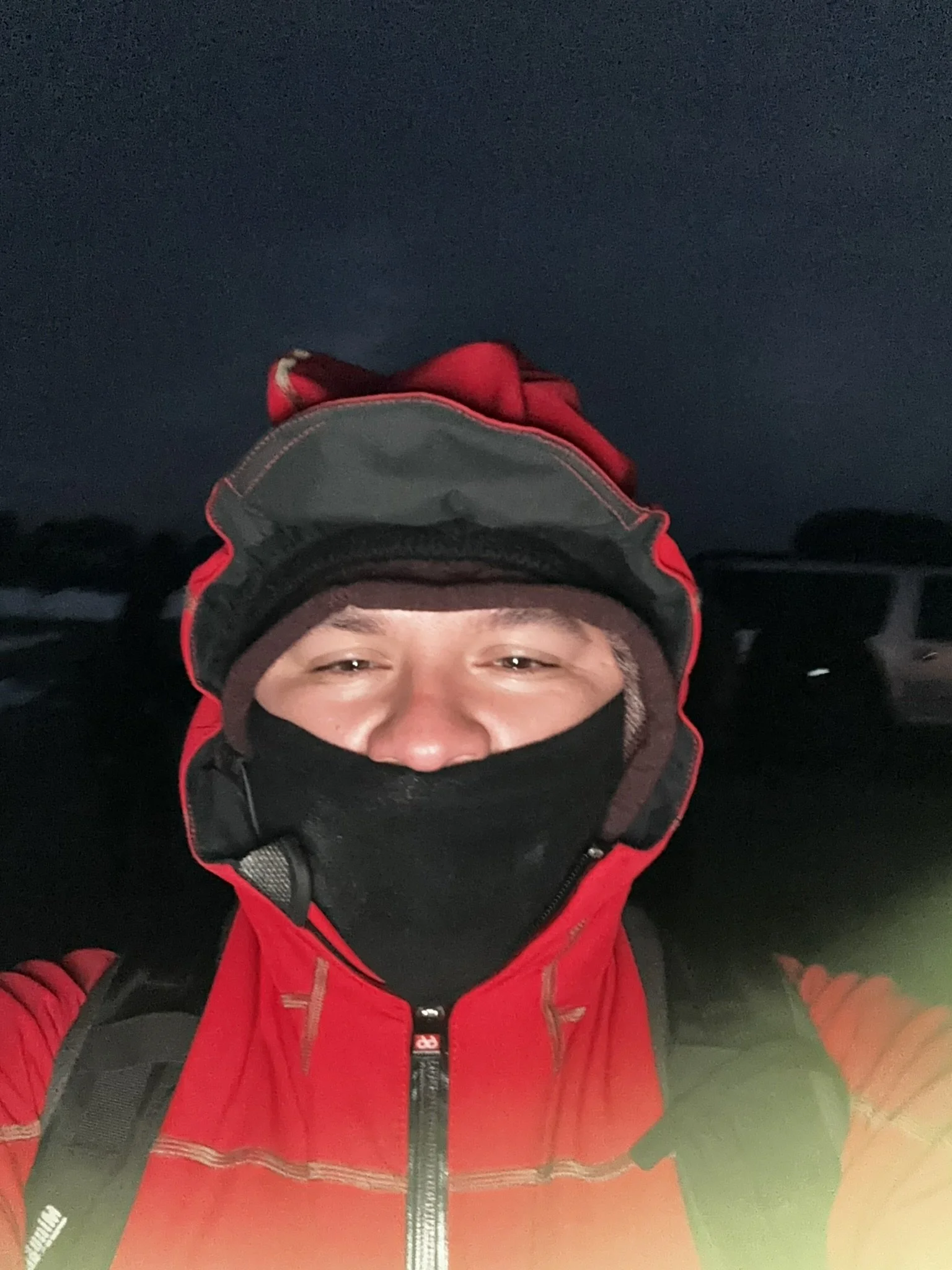 A person in outdoor gear wearing a red jacket, black face mask, and hood, standing outside at night.