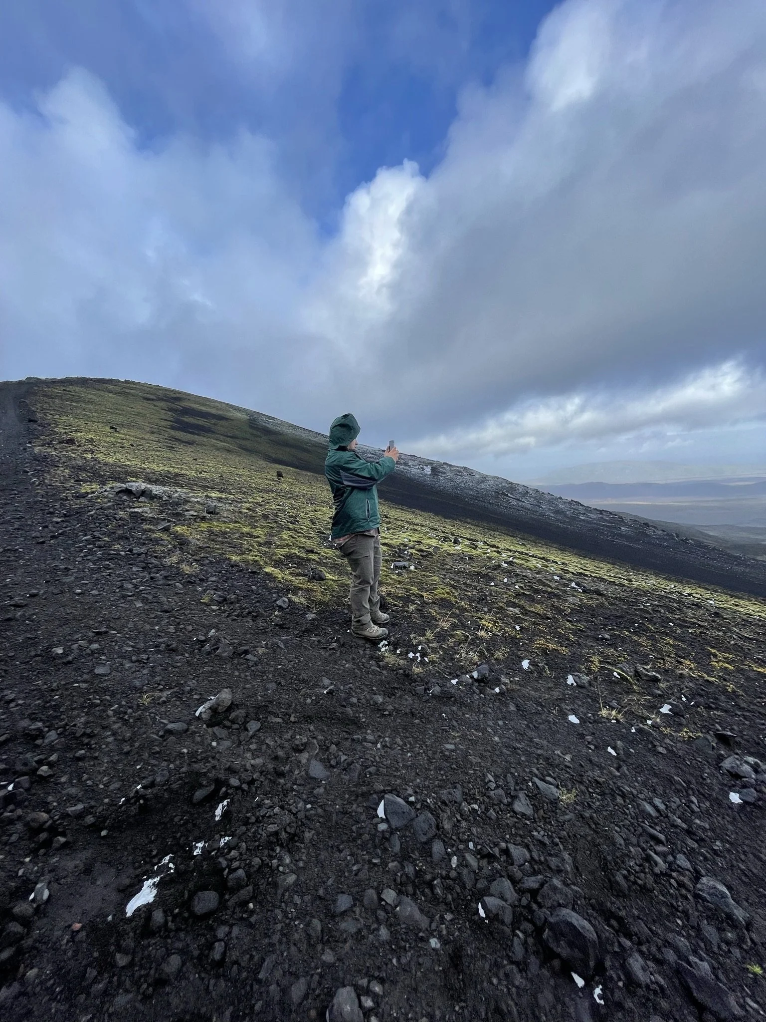 A person in a green jacket and gray pants is standing on a mountain slope, taking a photo with a smartphone. The slope is covered with dark volcanic rocks and patches of moss, with a cloudy sky overhead.