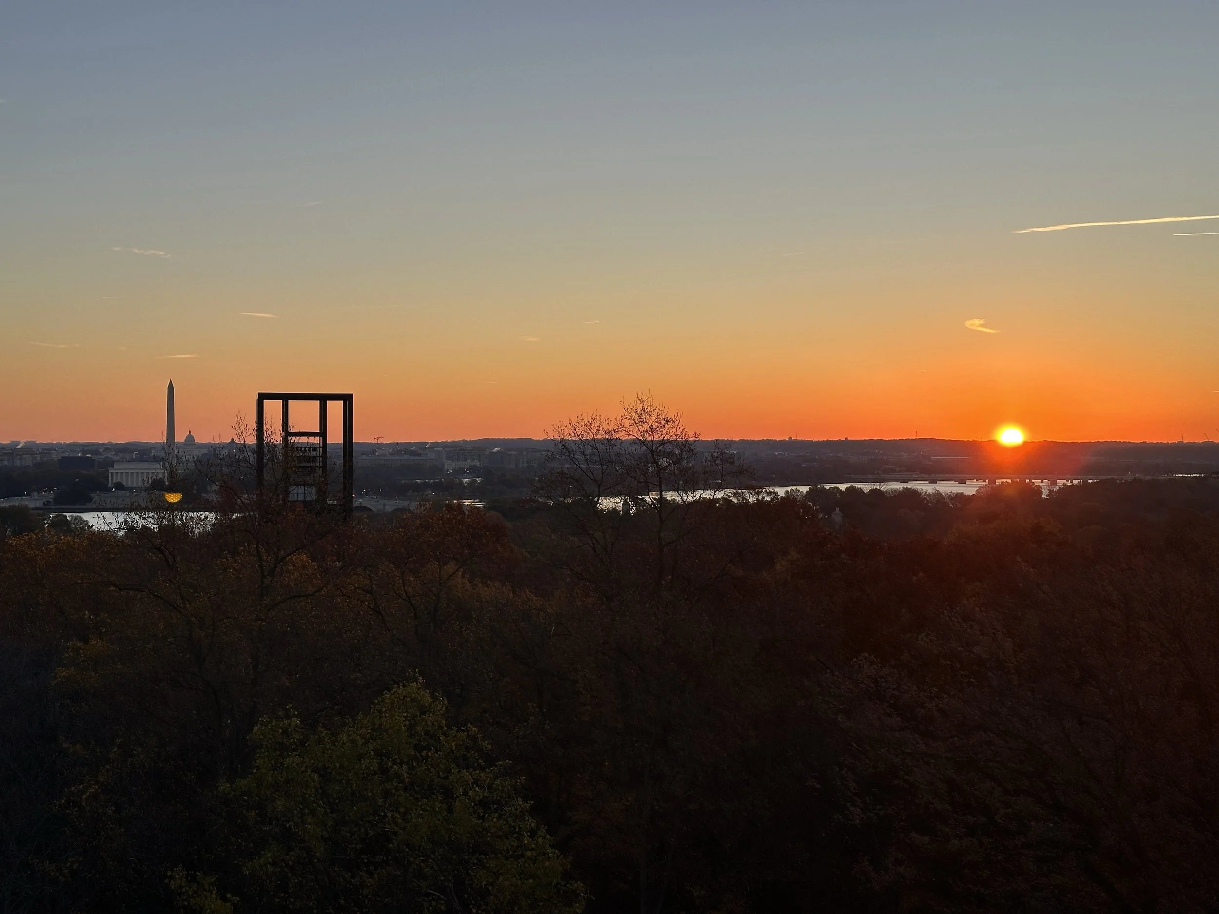 Sunset over Washington, D.C., with the Washington Monument and the U.S. Capitol in the distance, viewed from a park with trees in the foreground.