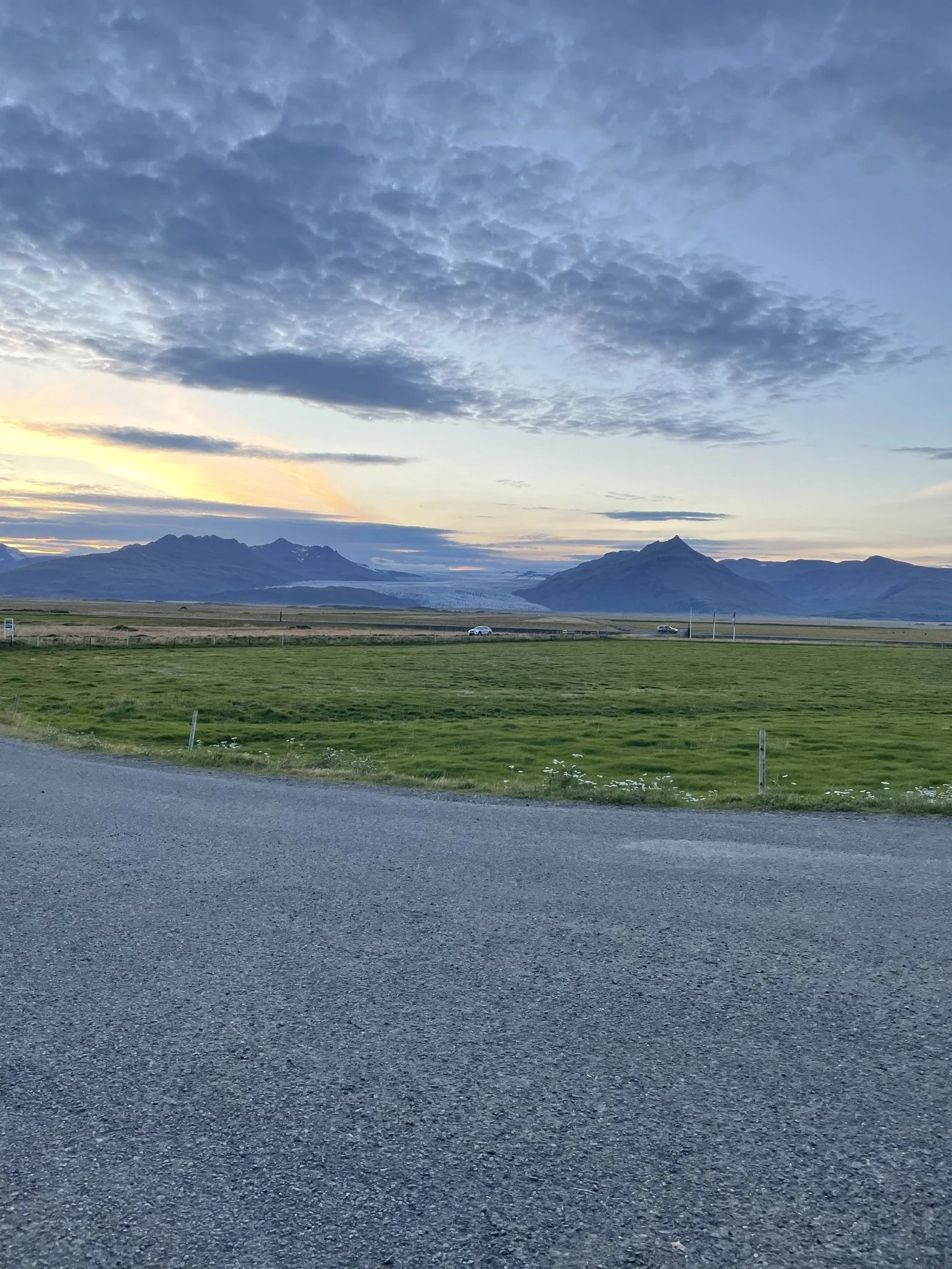Scenic landscape featuring a grassy field, a winding road, distant mountains, and a partly cloudy sky at sunset.