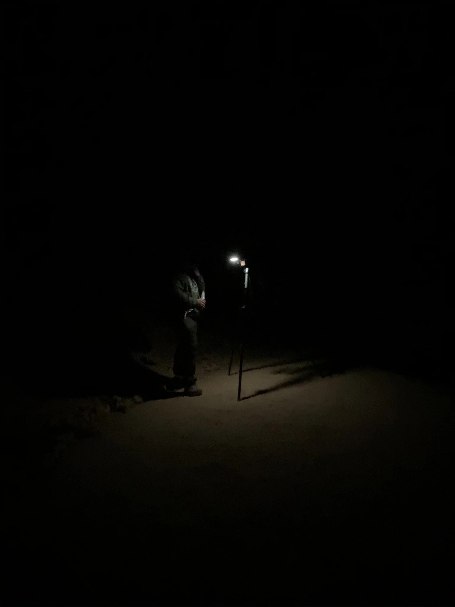 Person standing outdoors in the dark, illuminated by a small portable light, with a tripod nearby.