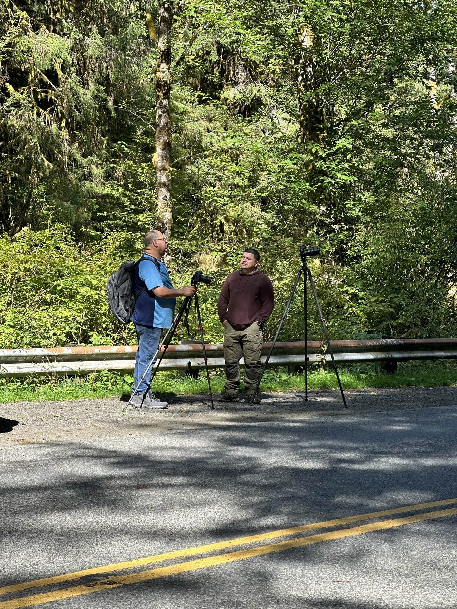 Two men standing beside a guardrail on a road, with one man holding a camera on a tripod and the other man standing nearby, in front of a dense wooded forest.