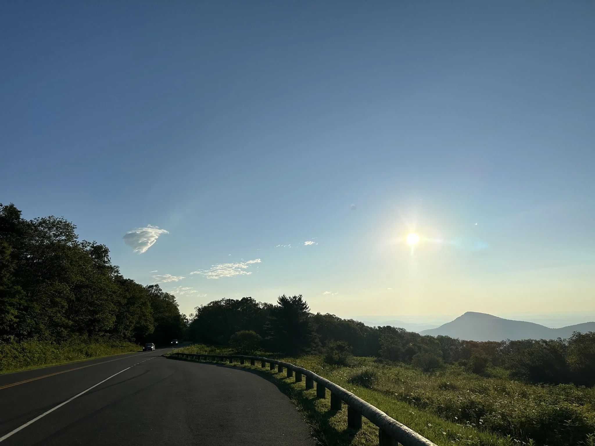 A winding road through a lush green mountainous landscape with a clear blue sky and bright sun.